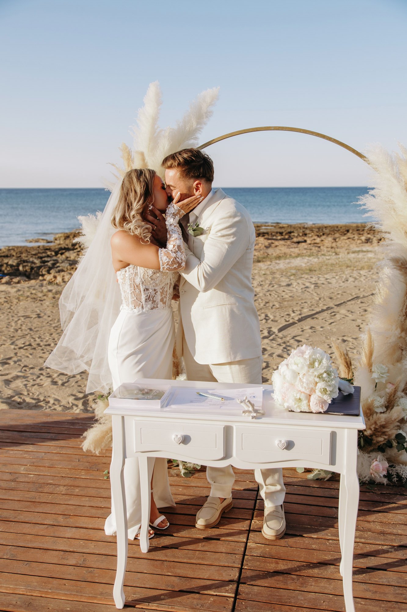 A couple in wedding attire sharing a kiss at an outdoor beach wedding ceremony with a heart-shaped floral arch and ocean view.