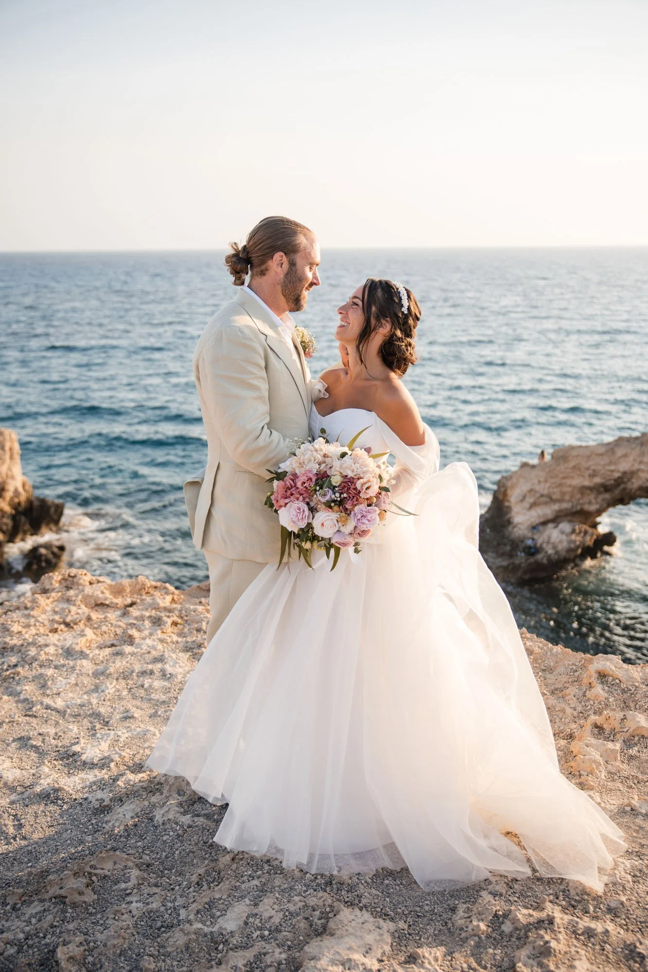 A newlywed couple on a rocky shoreline during sunset, dressed in wedding attire, gazing at each other and smiling, with the ocean in the background.