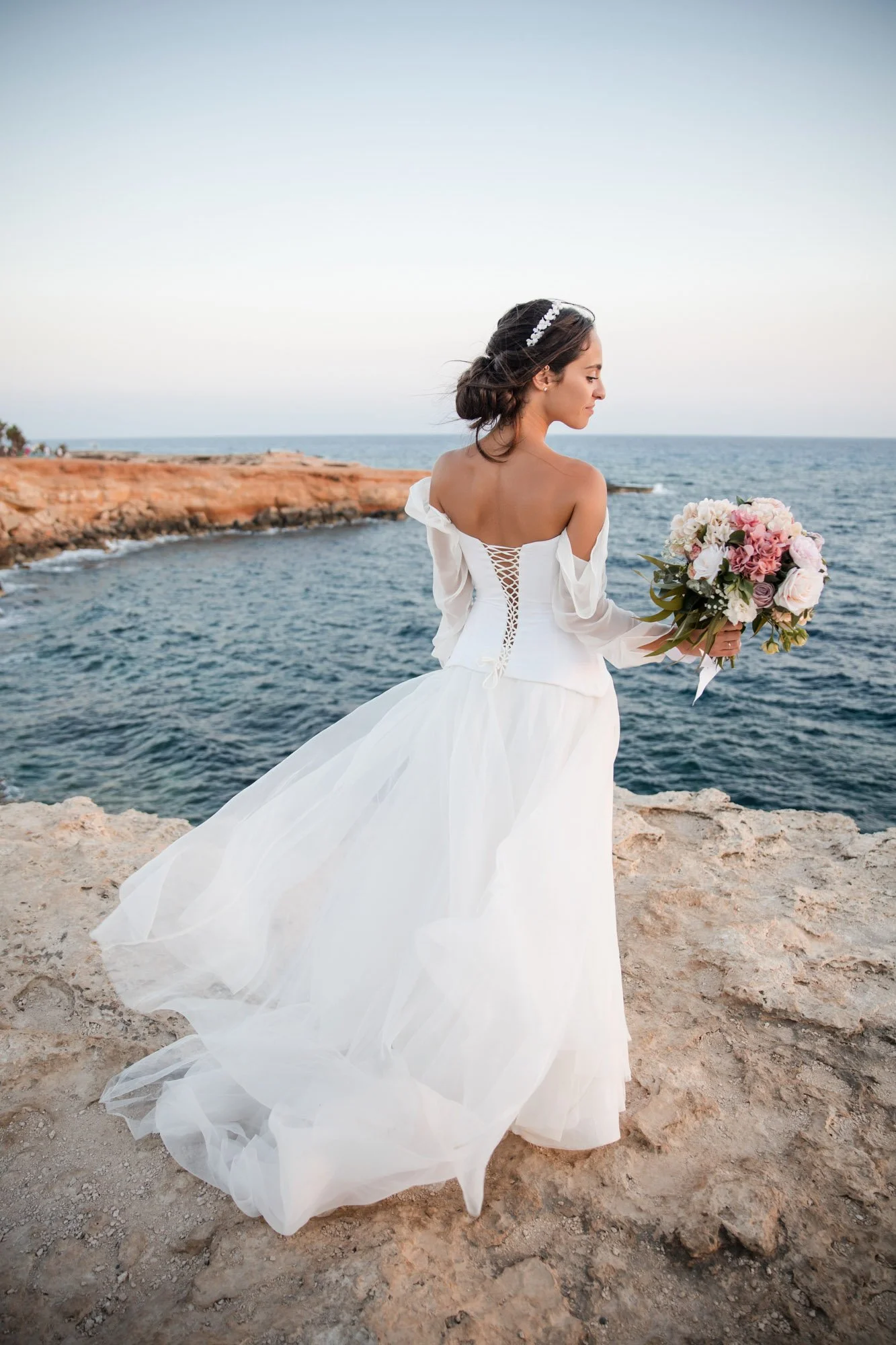 A bride in a white wedding dress holding a bouquet of pink and white flowers stands on a rocky cliff overlooking the ocean during sunset.
