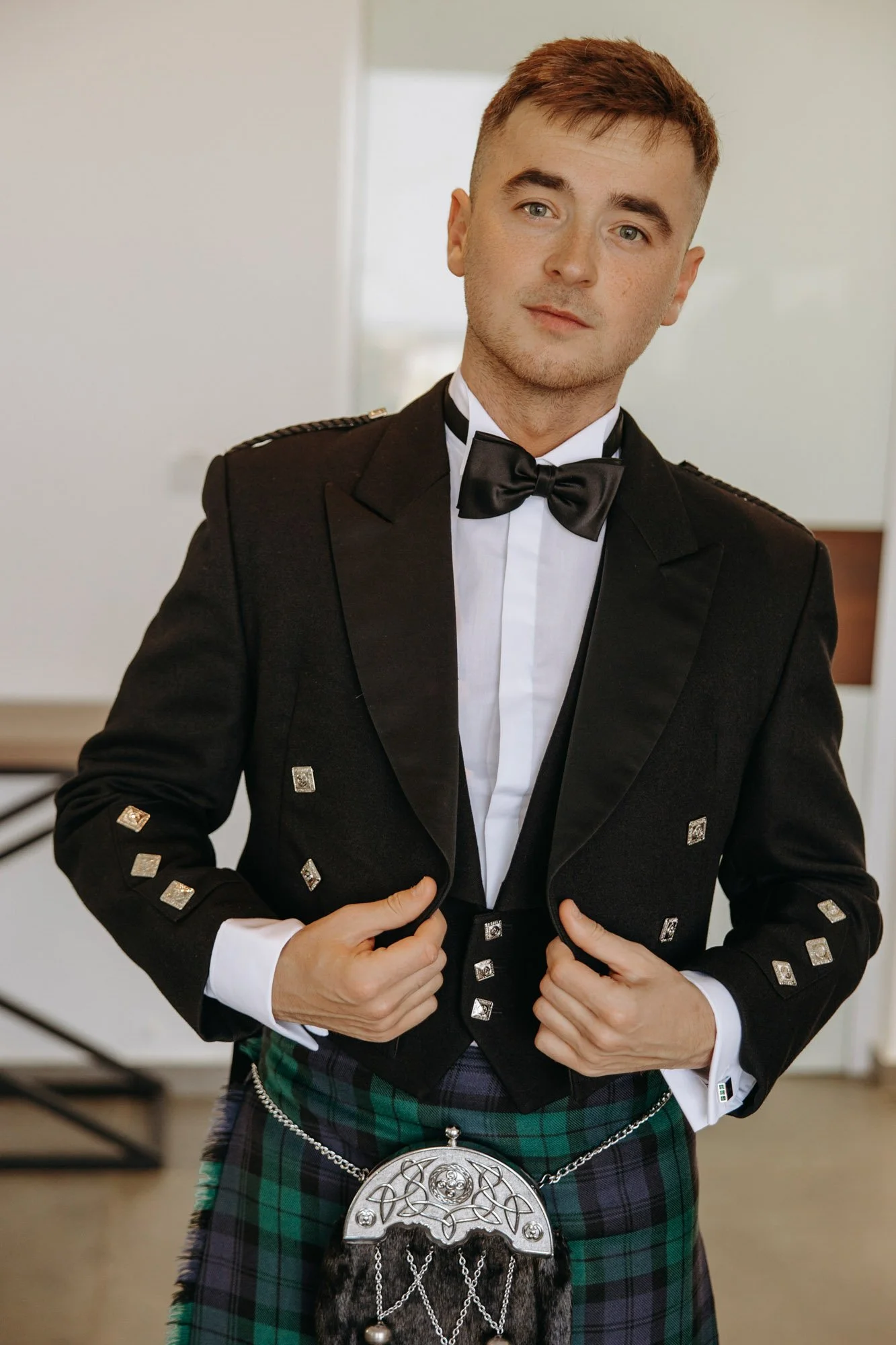 Young man in formal attire wearing a black jacket with silver buttons, a white shirt, black bow tie, and a traditional Scottish kilt with a sporran, standing indoors.
