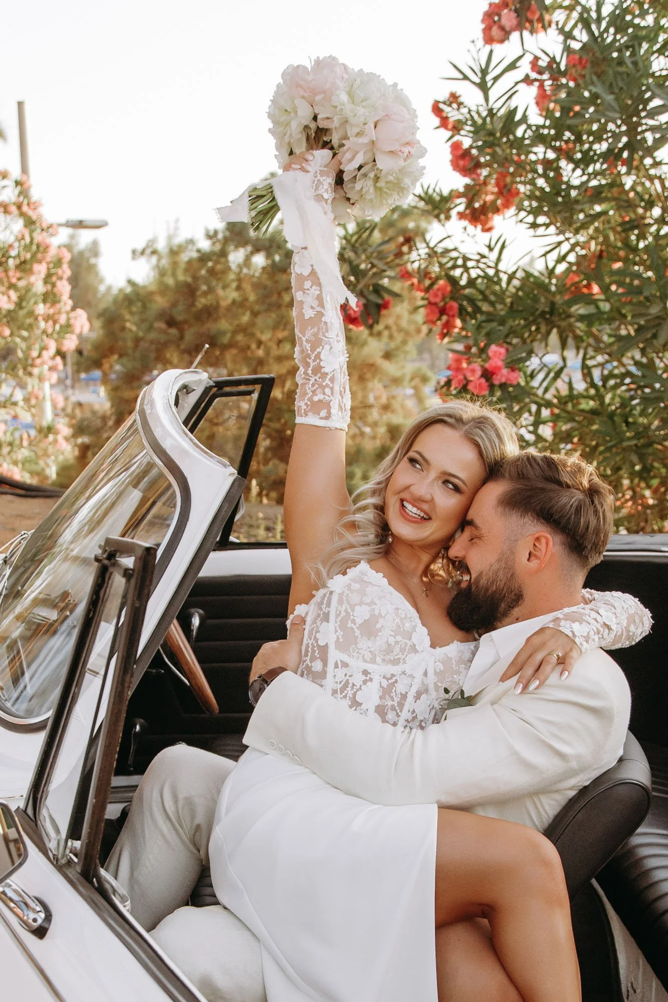 A bride and groom sitting in a vintage convertible car, embracing and smiling. The bride is holding a bouquet of flowers above her head. The background has trees and pink flowers.