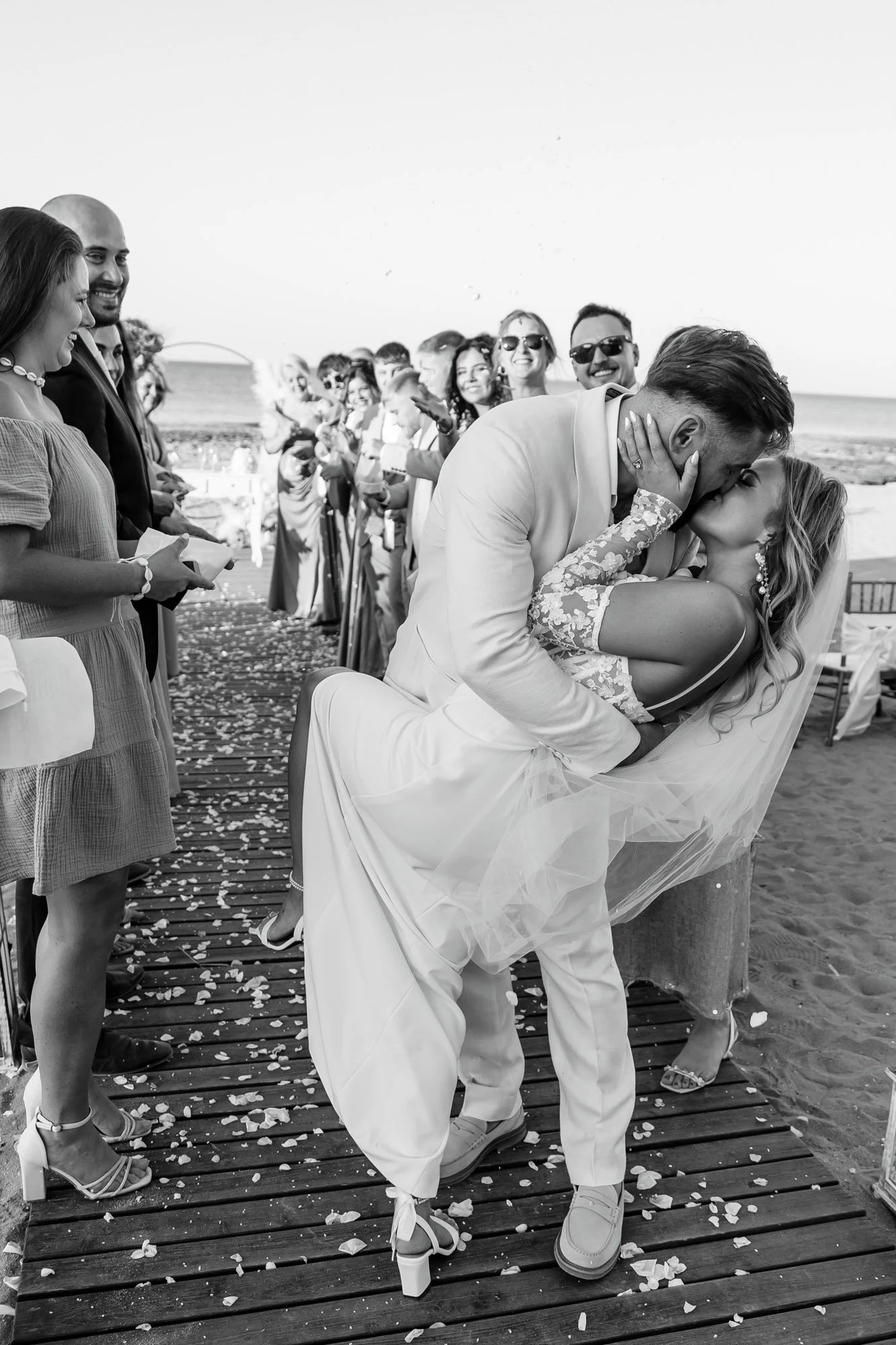 A couple, the groom in a white suit and the bride in a wedding dress, sharing a kiss during their beach wedding ceremony while holding each other and leaning on each other on a wooden pier, with guests in the background watching and smiling.