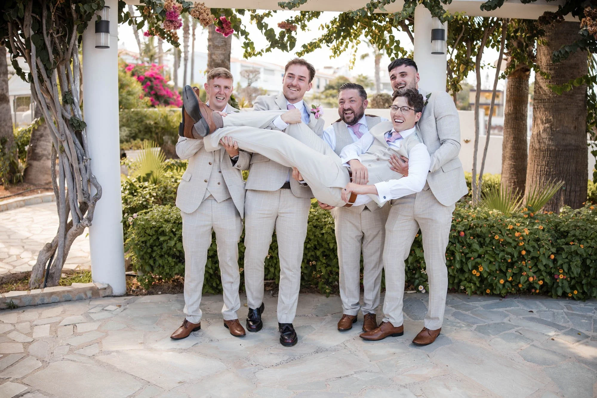 Group of six men in beige suits lifting a man in a beige suit and white shirt in a wedding setting outdoors, surrounded by trees and colorful flowers, smiling and laughing.