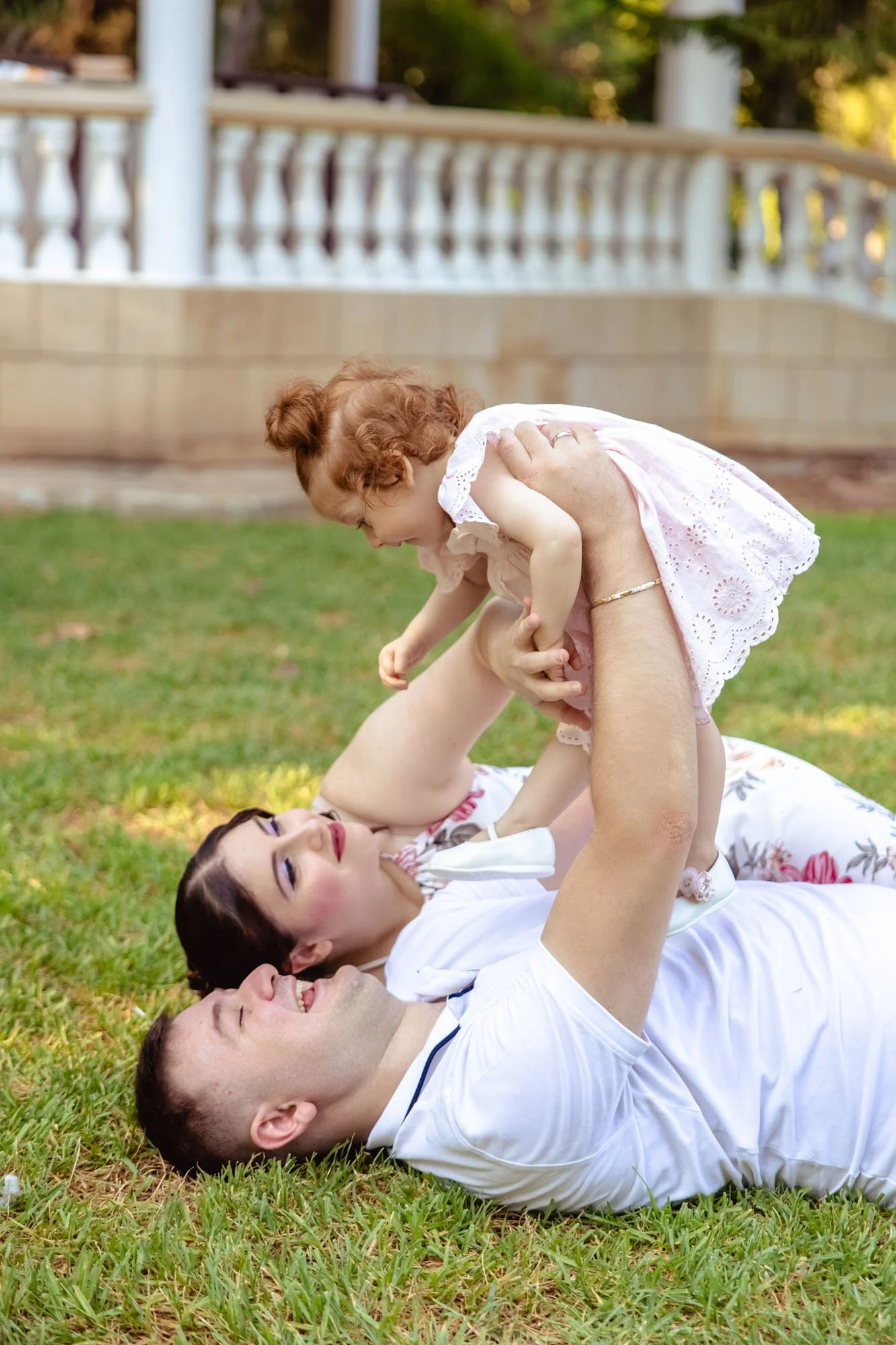 A family of three lying on grass outdoors, with the father holding a young girl above him. They are smiling and enjoying time together in a park or garden setting.