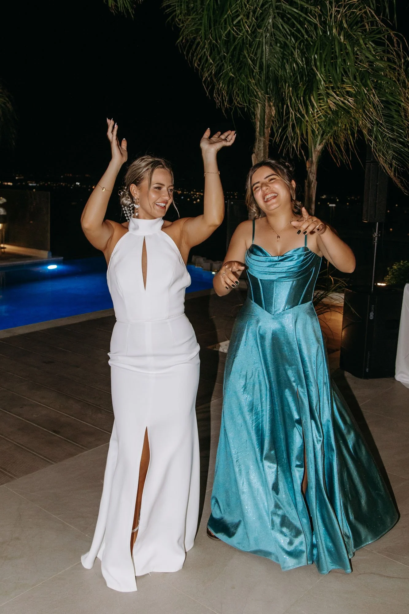 Two women dancing and smiling at an outdoor party at night, dressed in elegant evening gowns, by a swimming pool and palm trees.