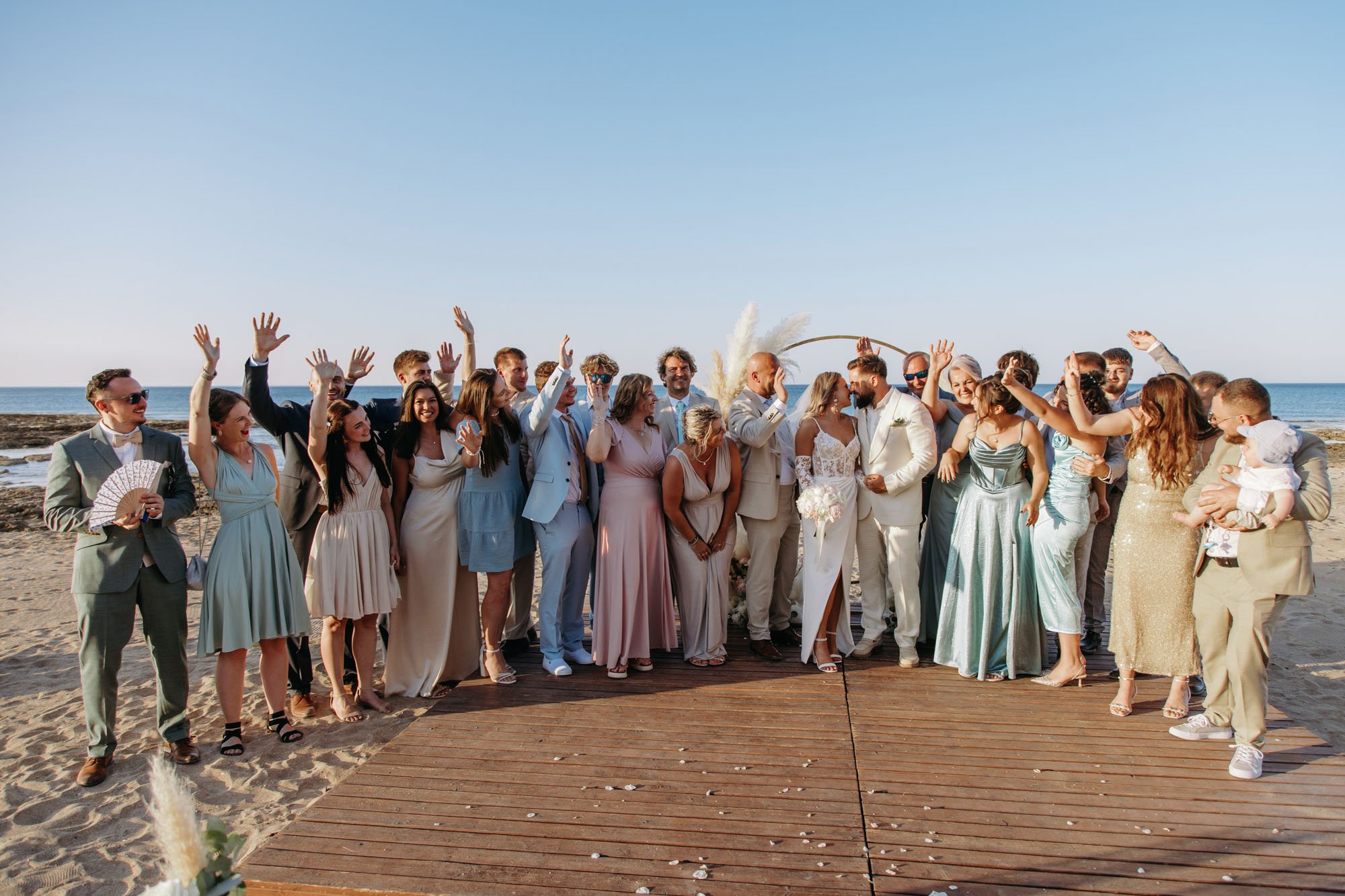 Group of people dressed in formal attire on a beach, celebrating a wedding with the ocean in the background.