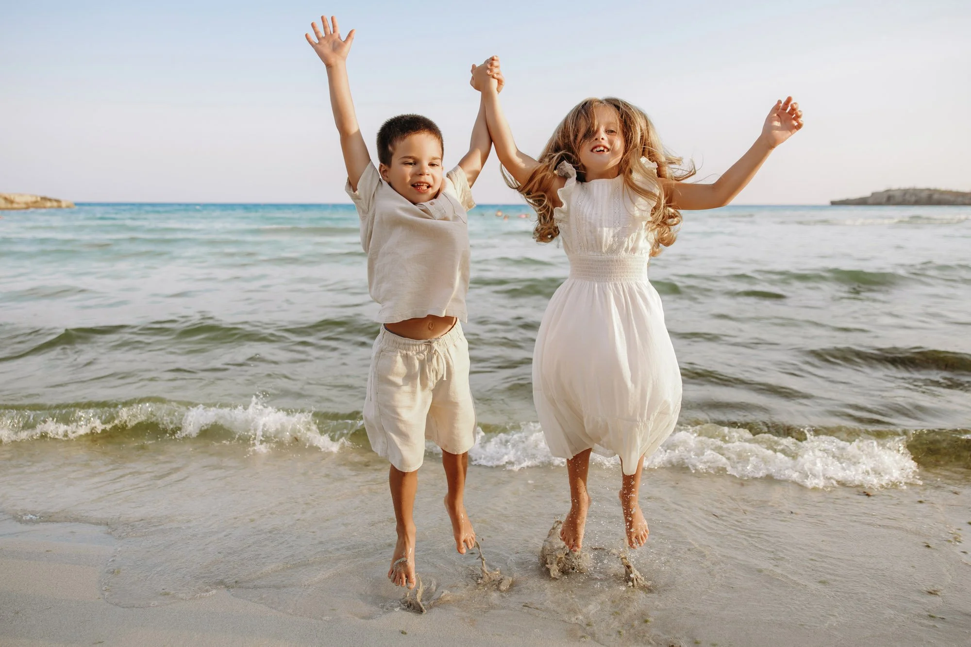 Two children, a boy and a girl, holding hands and jumping in the shallow water of a beach, smiling, with waves and a clear sky in the background.