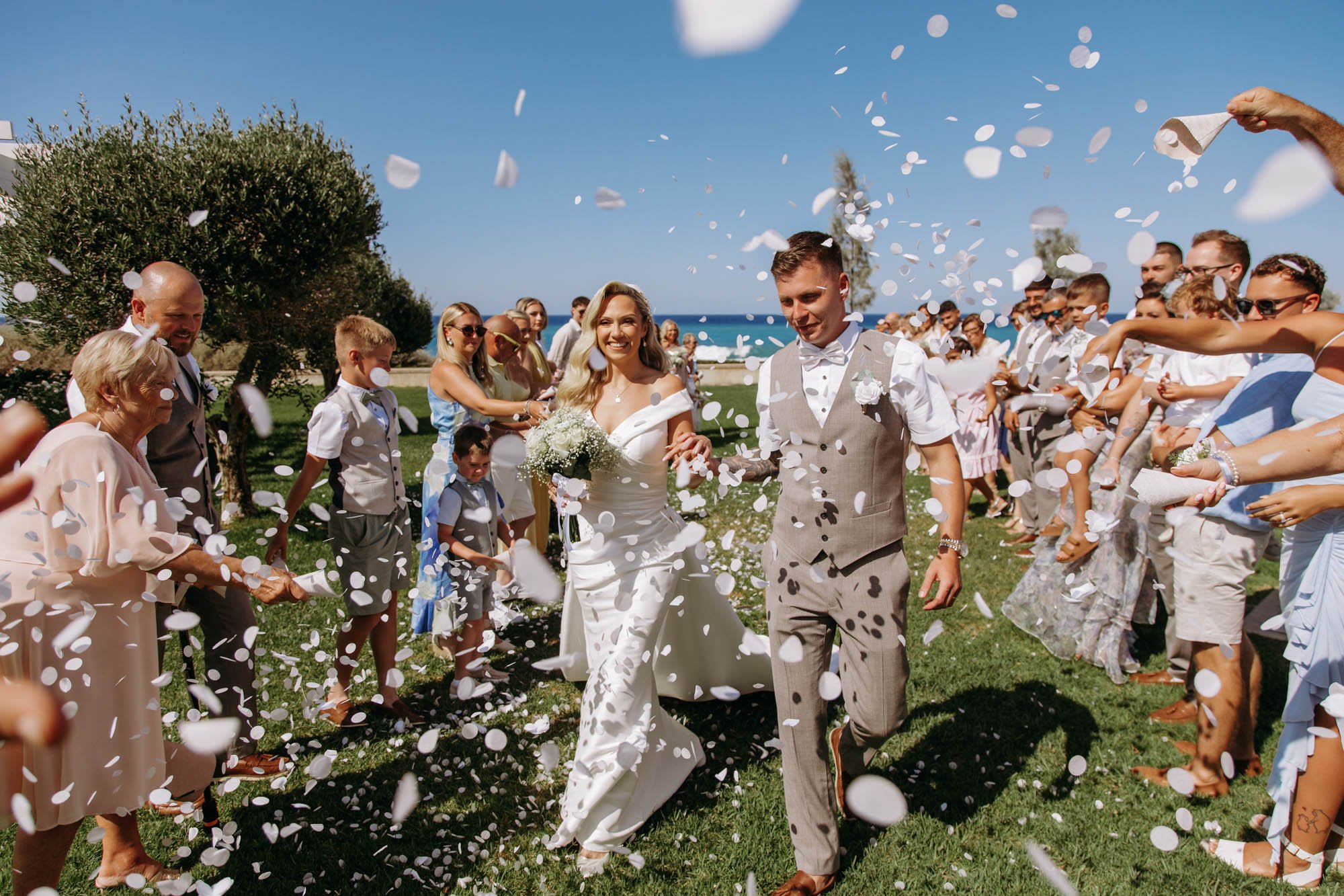 Bride and groom walking through confetti while wedding guests celebrate outdoors under a clear blue sky.