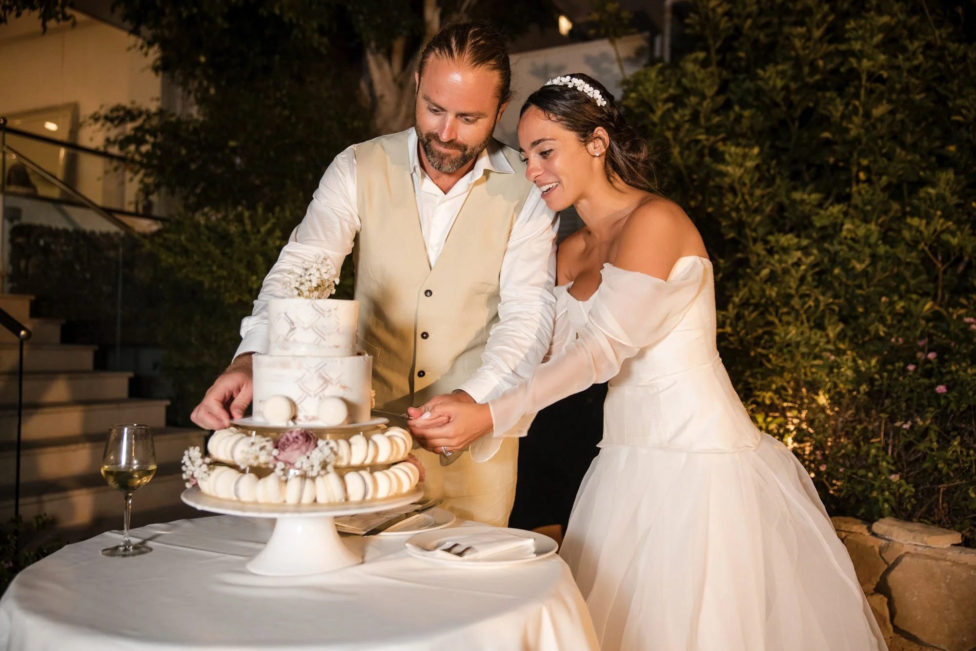 A bride and groom cut their wedding cake together at an outdoor reception, with the bride wearing a white off-the-shoulder gown and the groom in a cream vest and white shirt, surrounded by greenery and warm lighting.