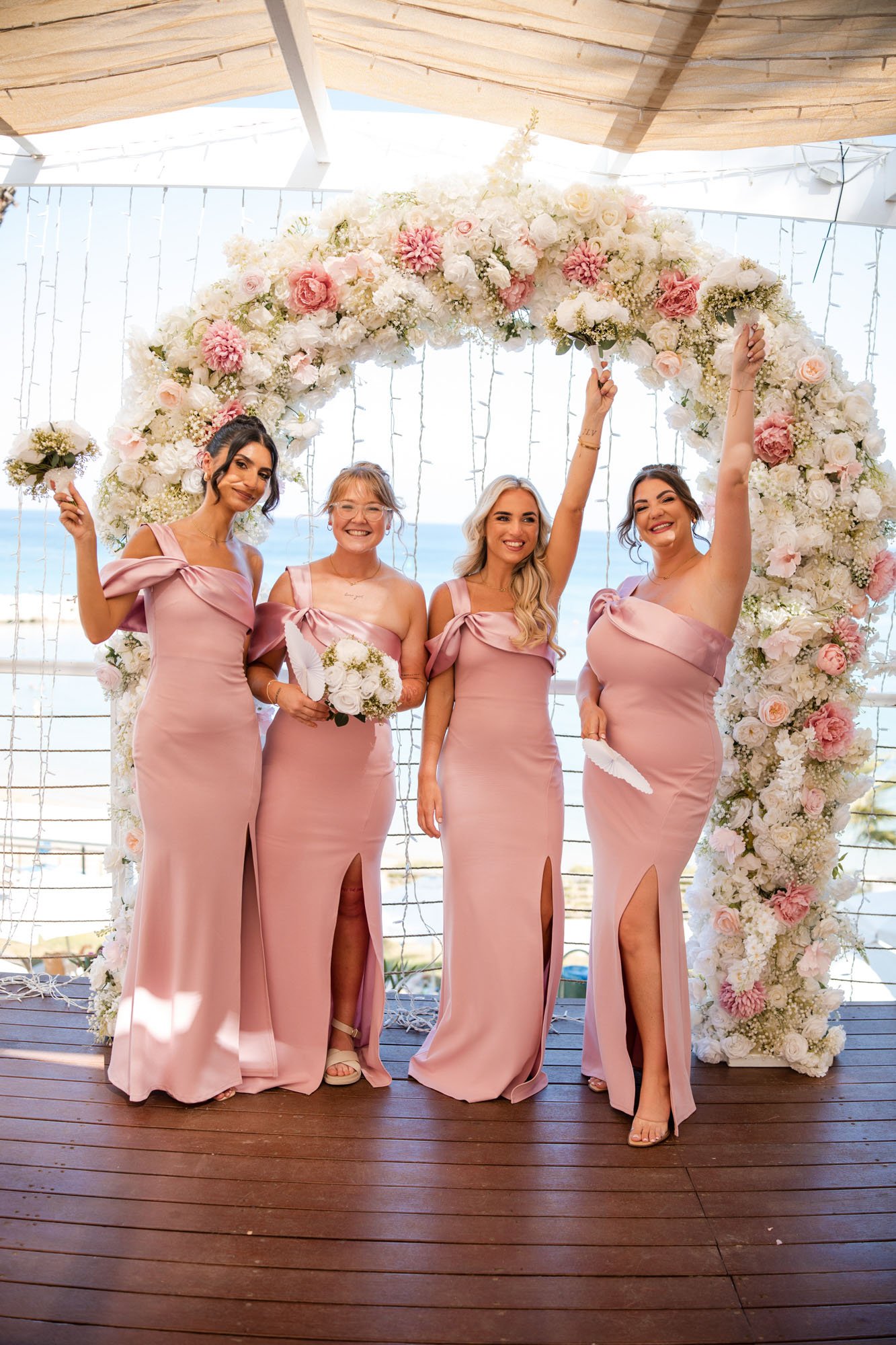 Four women in pink dresses standing under a floral arch during a wedding ceremony, with an ocean view in the background.