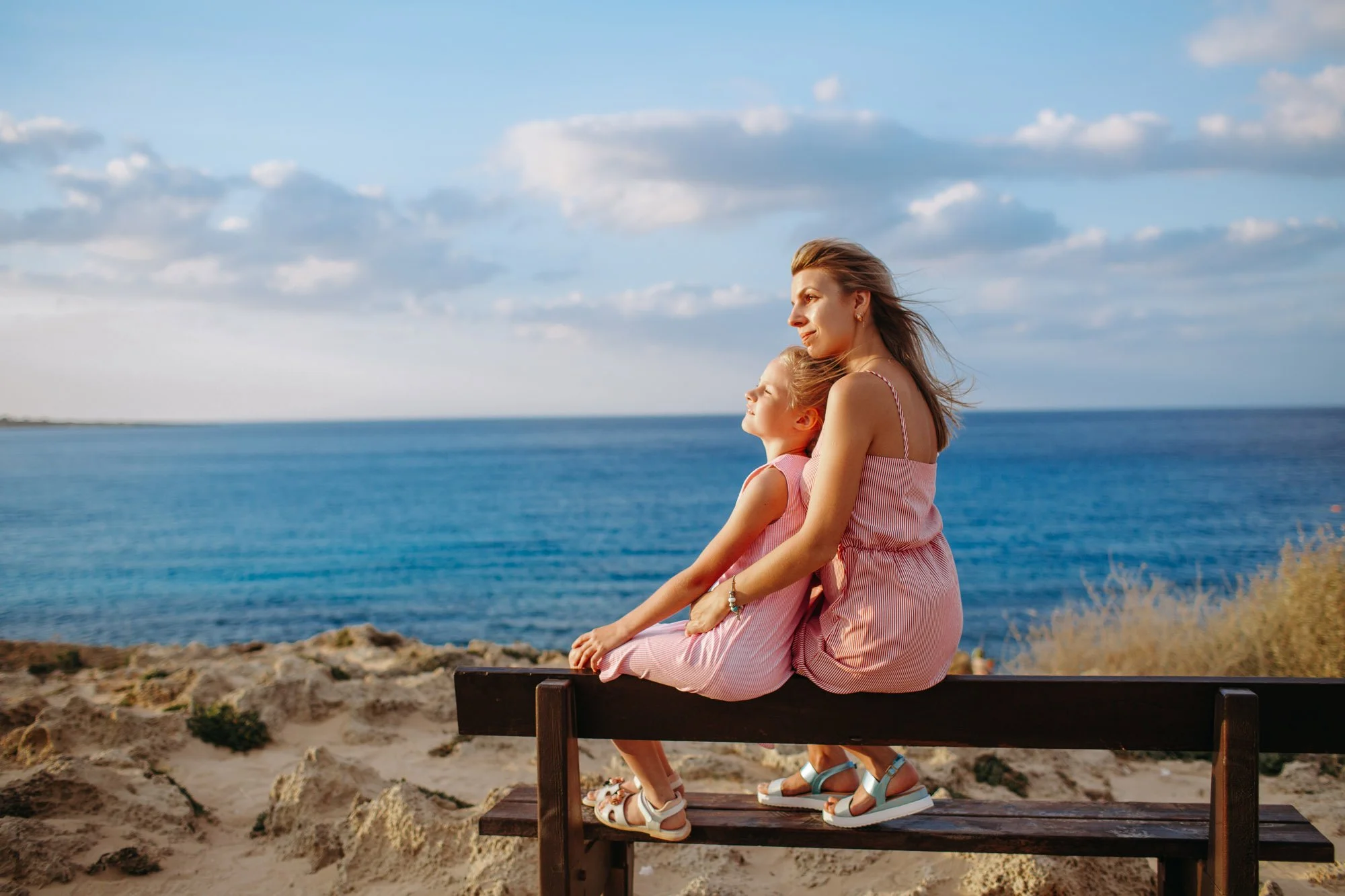 A woman and a young girl sit together on a wooden bench by the ocean on a partly cloudy day, enjoying the view and airy breeze.