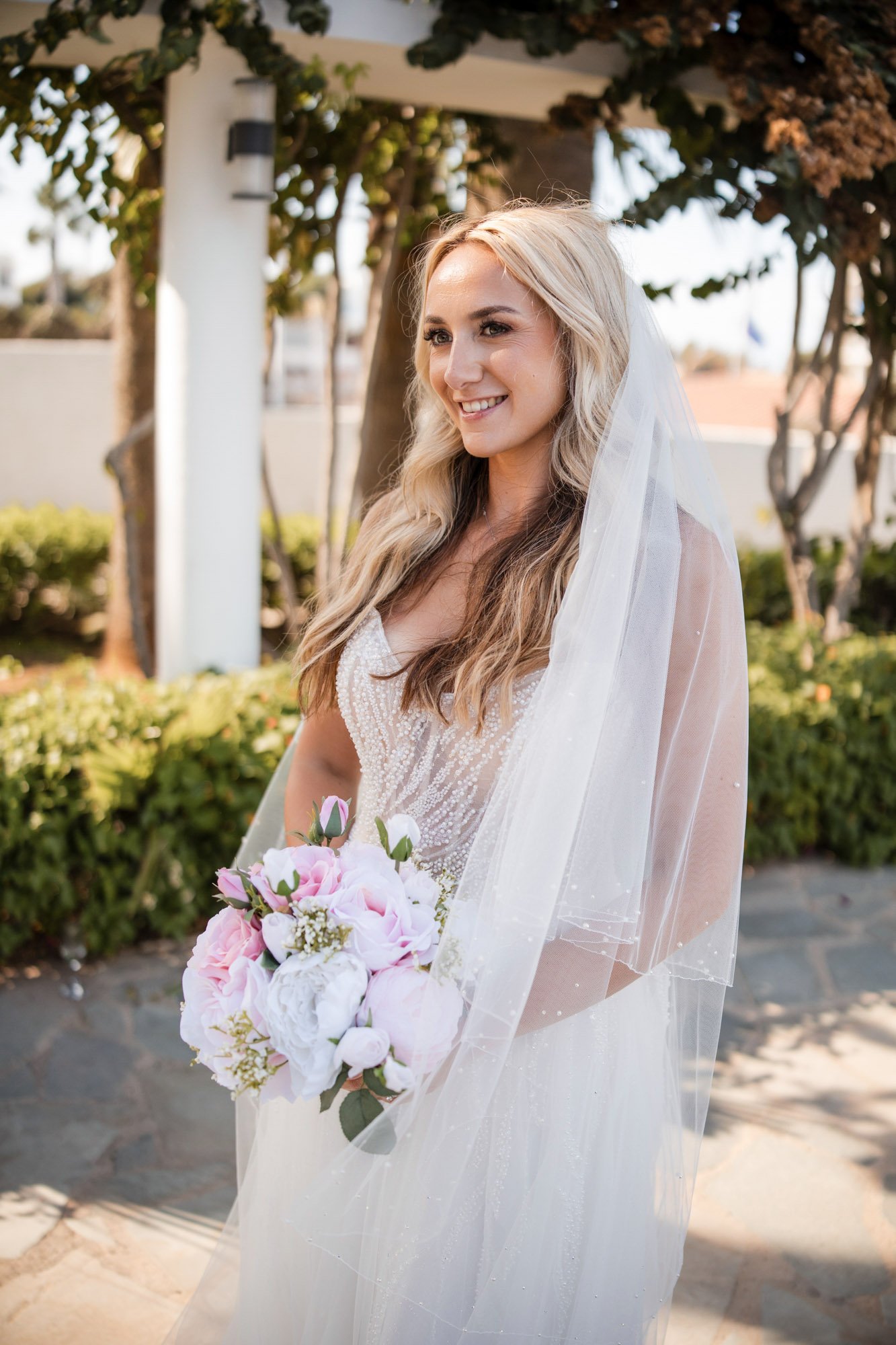 Wedding Photographer Golden Coast, Protaras A smiling woman in a wedding dress holding a bouquet of pink and white flowers outdoors, with trees and bushes in the background.