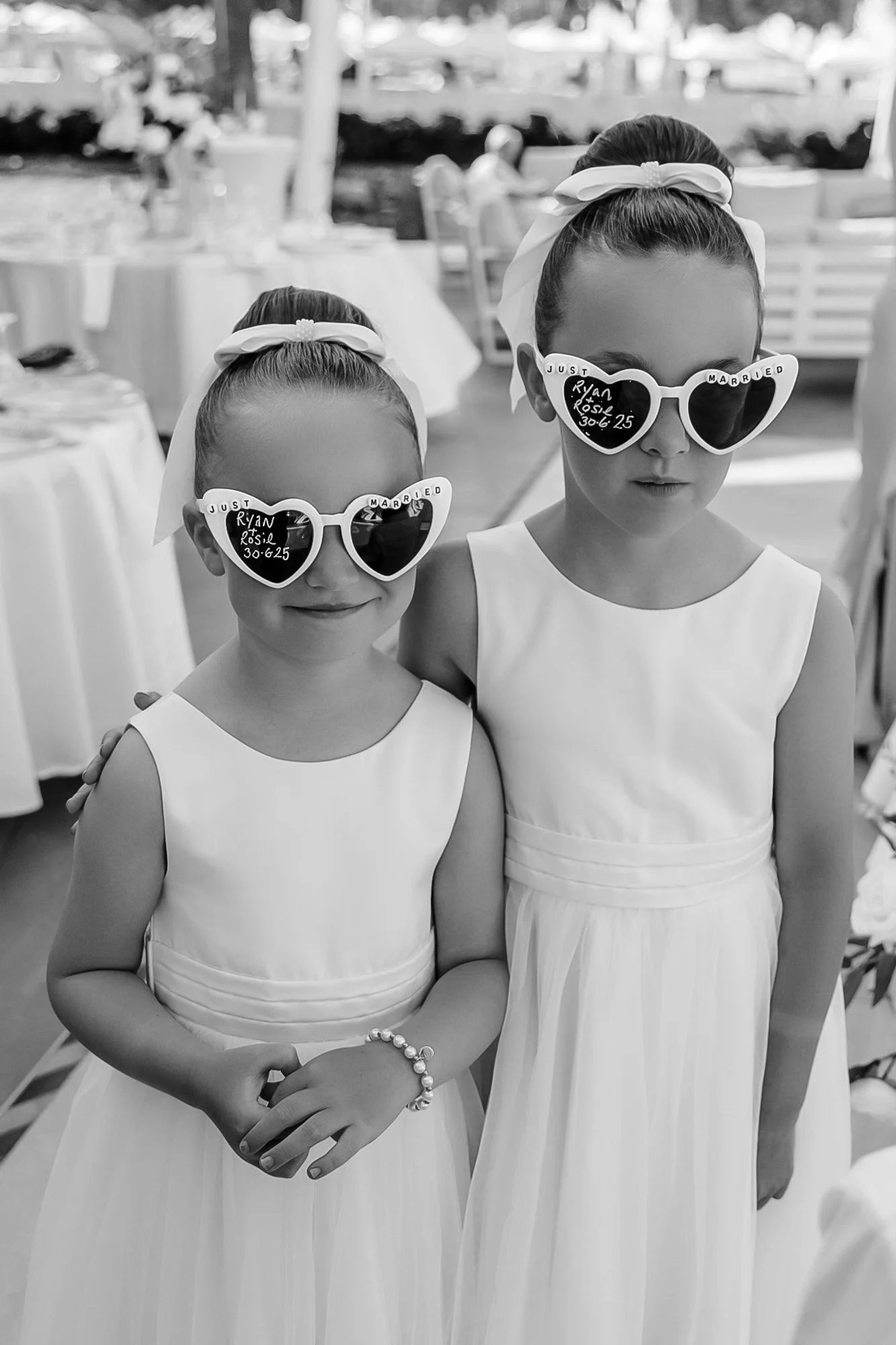 Two young girls dressed in white dresses wearing sunglasses with decorations, posing together at a wedding.