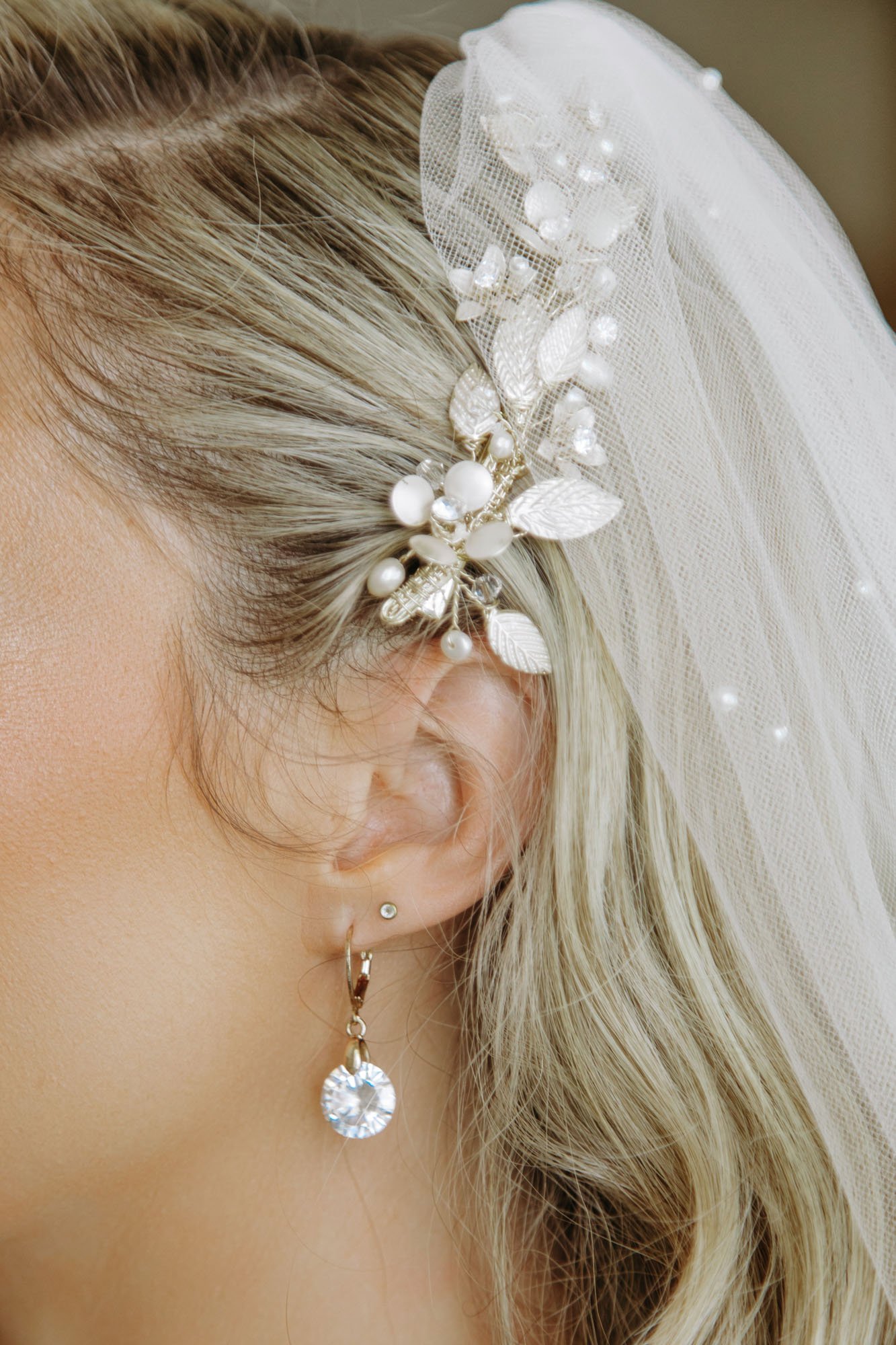 Close-up of a woman's ear with multiple earrings, including a dangling crystal earring and a pearl earring, with a decorative hairpiece featuring white pearls and leaf-shaped embellishments, and a sheer veil with small pearls.