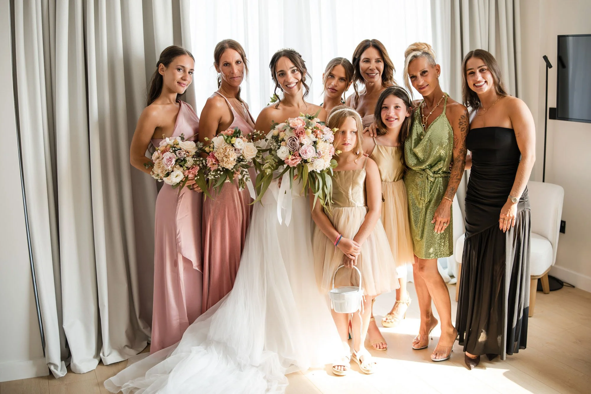 A group of women and girls in a bridal party, posing together indoors. The bride is holding a bouquet, wearing a white wedding gown, surrounded by bridesmaids and flower girls in pastel and metallic dresses.
