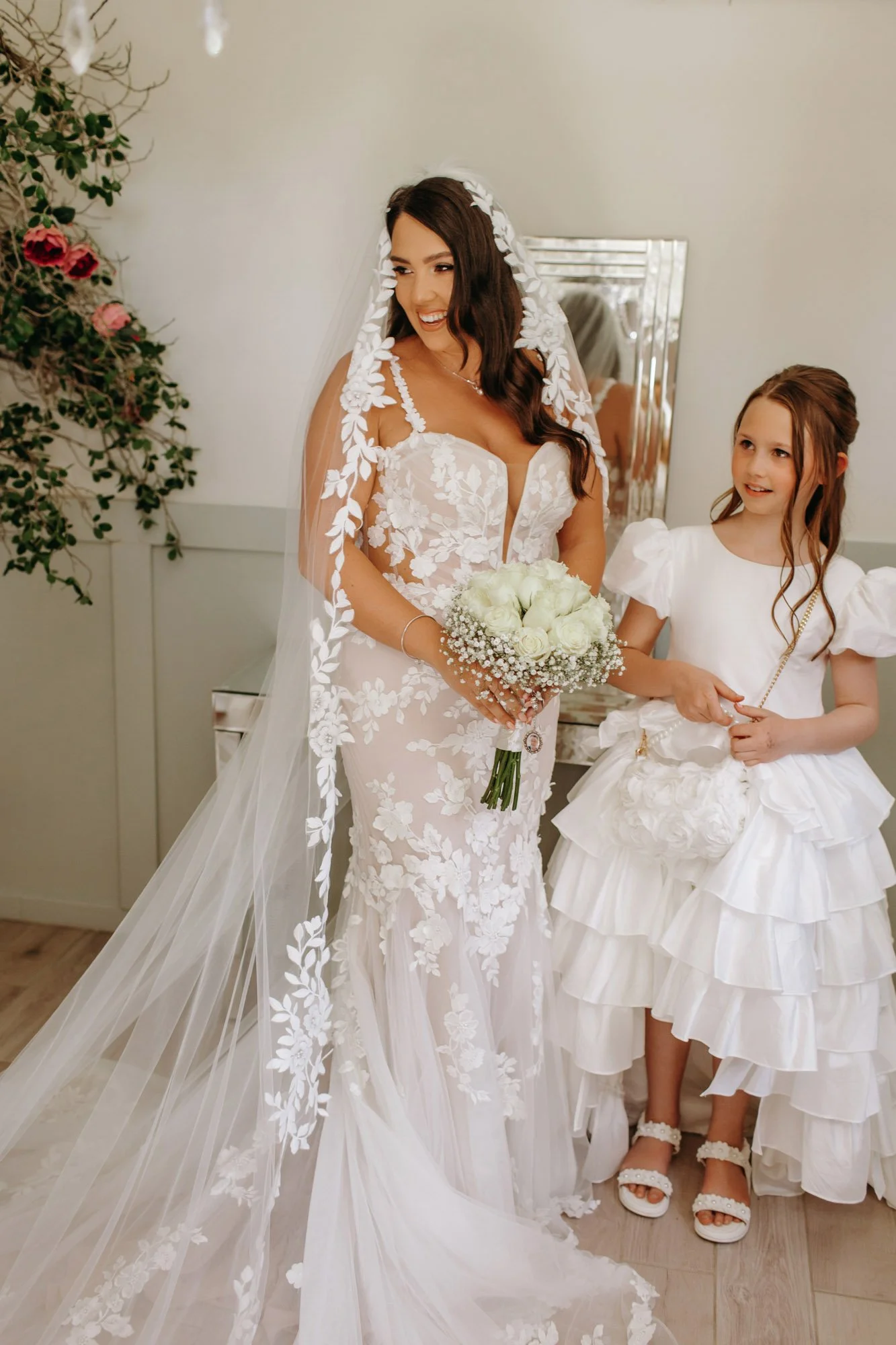 A bride in a white wedding dress with floral lace details, holding a bouquet of white flowers, standing beside a young girl in a white dress with ruffled layers, inside a decorated room.