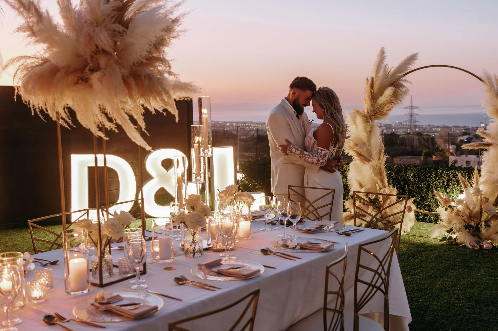 A couple in wedding attire embracing at an outdoor celebration table during sunset, decorated with candles, flowers, and pampas grass.