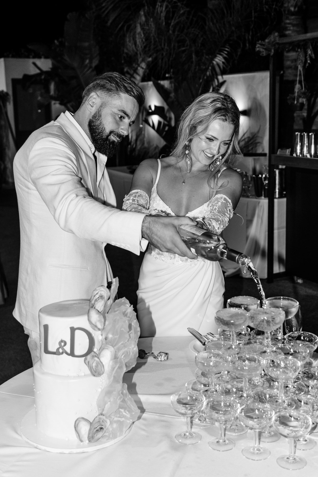 A black and white photo of a bride and groom pouring champagne into a pyramid of glasses at their wedding reception, with a wedding cake with 