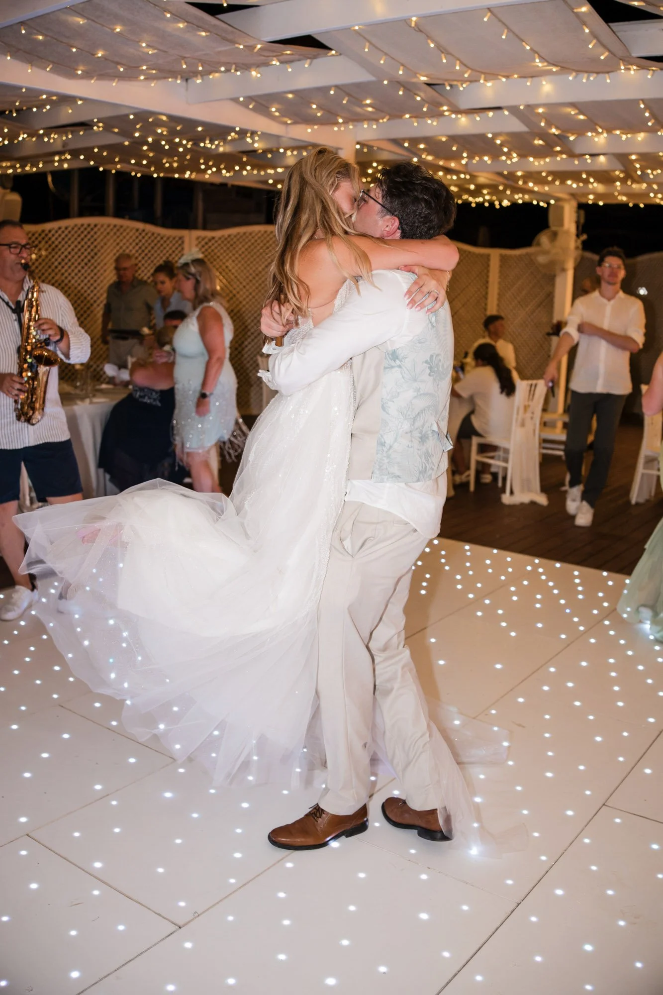 A bride and groom share a dance at their wedding reception, surrounded by guests and string lights.