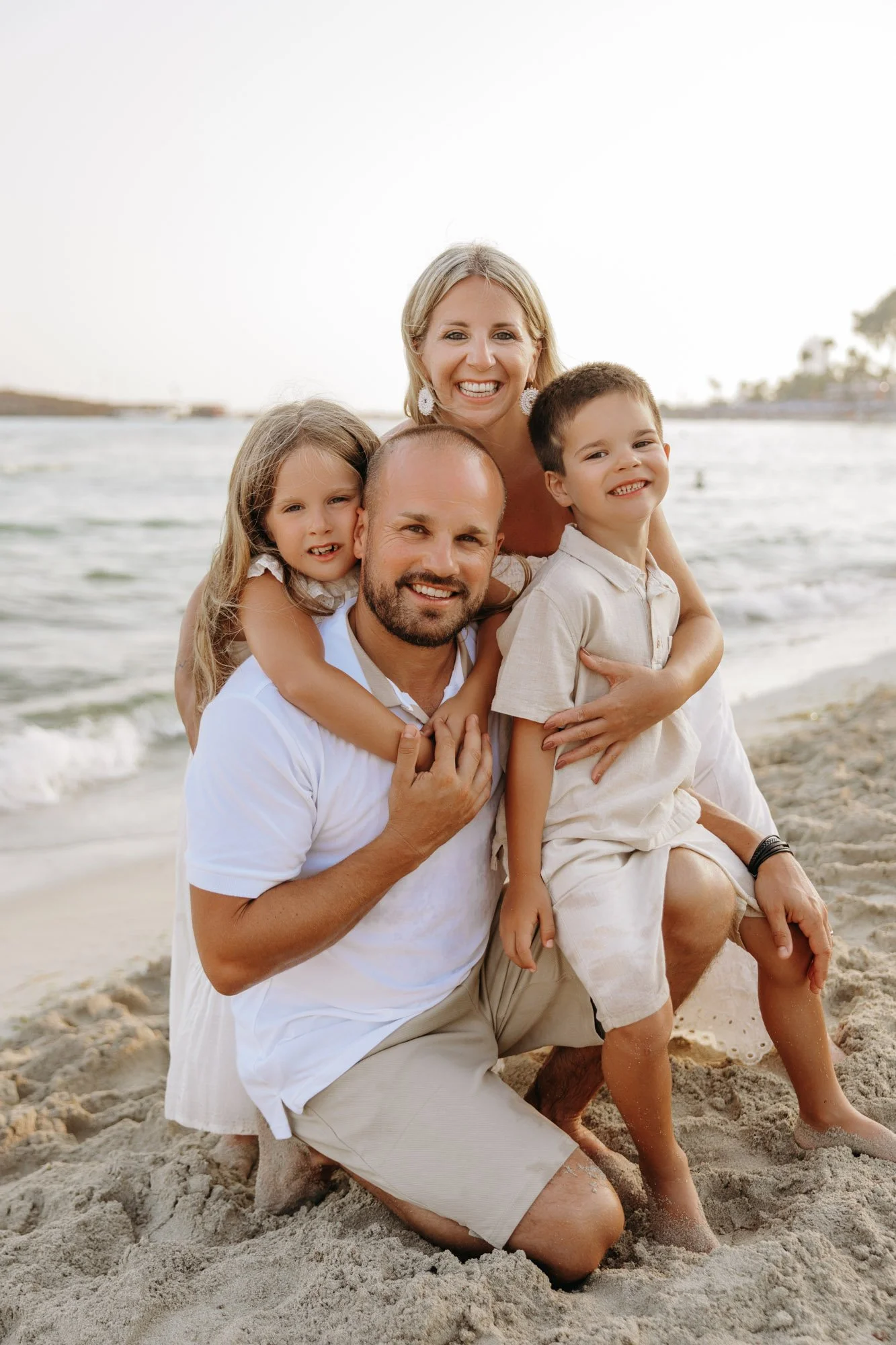 A happy family of four at the beach, with ocean and palm trees in the background, smiling and embracing each other.