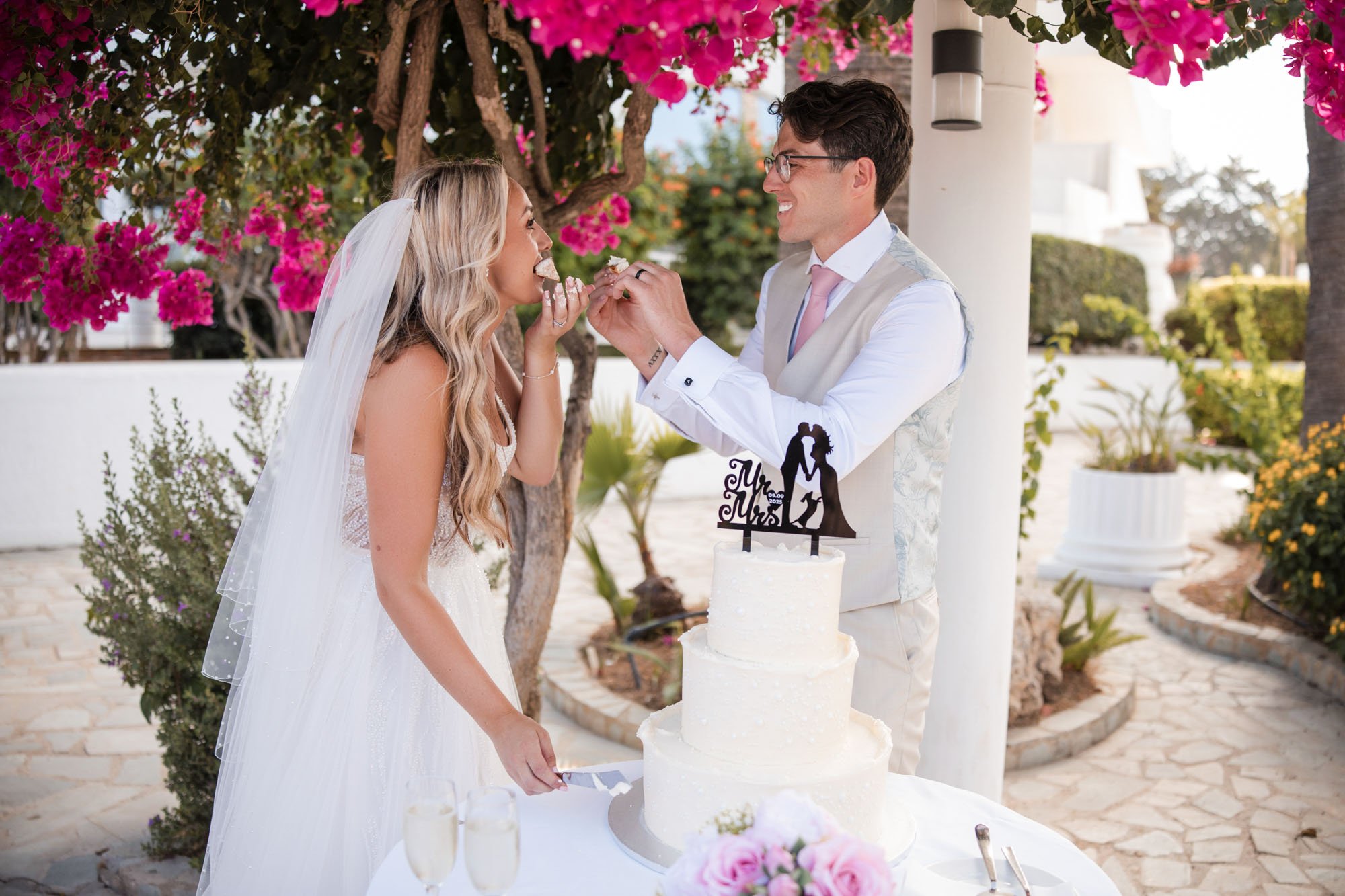 Bride and groom celebrating their wedding outdoors, with the groom feeding the bride cake. There is a three-tier white wedding cake with a topper that says 'Mr. & Mrs.' The scene is decorated with pink flowers and greenery.