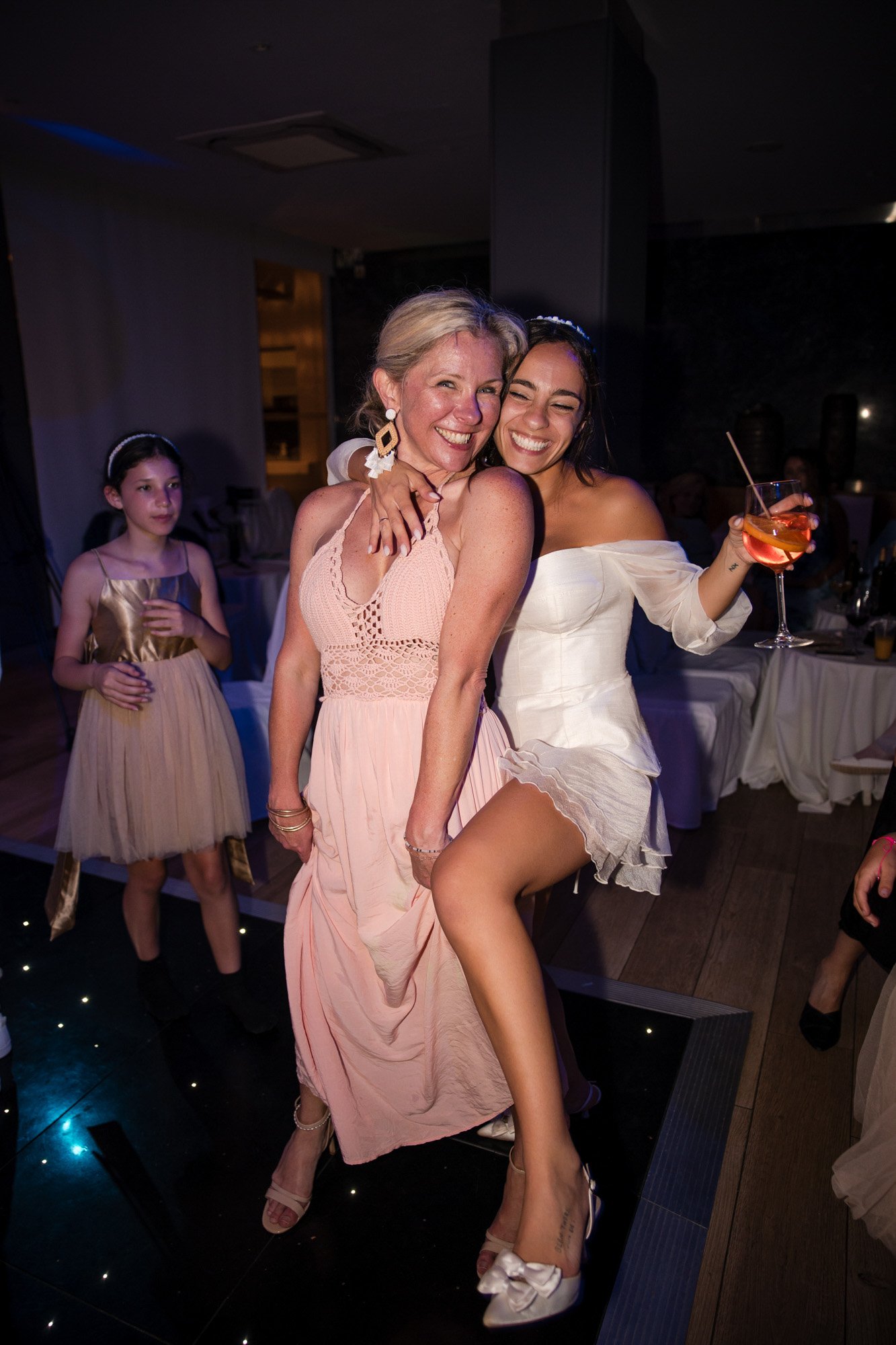 Two women celebrating at a party, smiling and hugging, one holding a drink, with a young girl in a gold dress in the background.