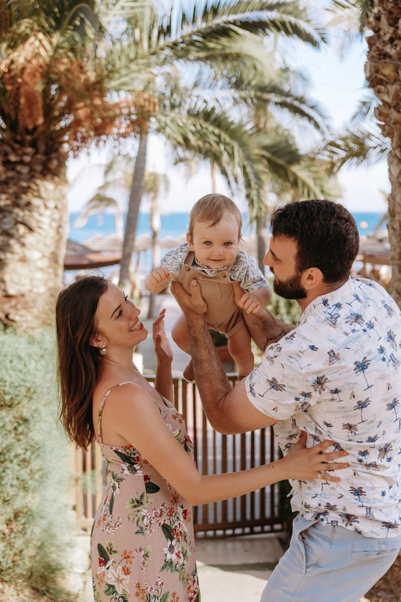 A family of three enjoying a day at the beach, with the father holding the baby in front of palm trees and ocean background.