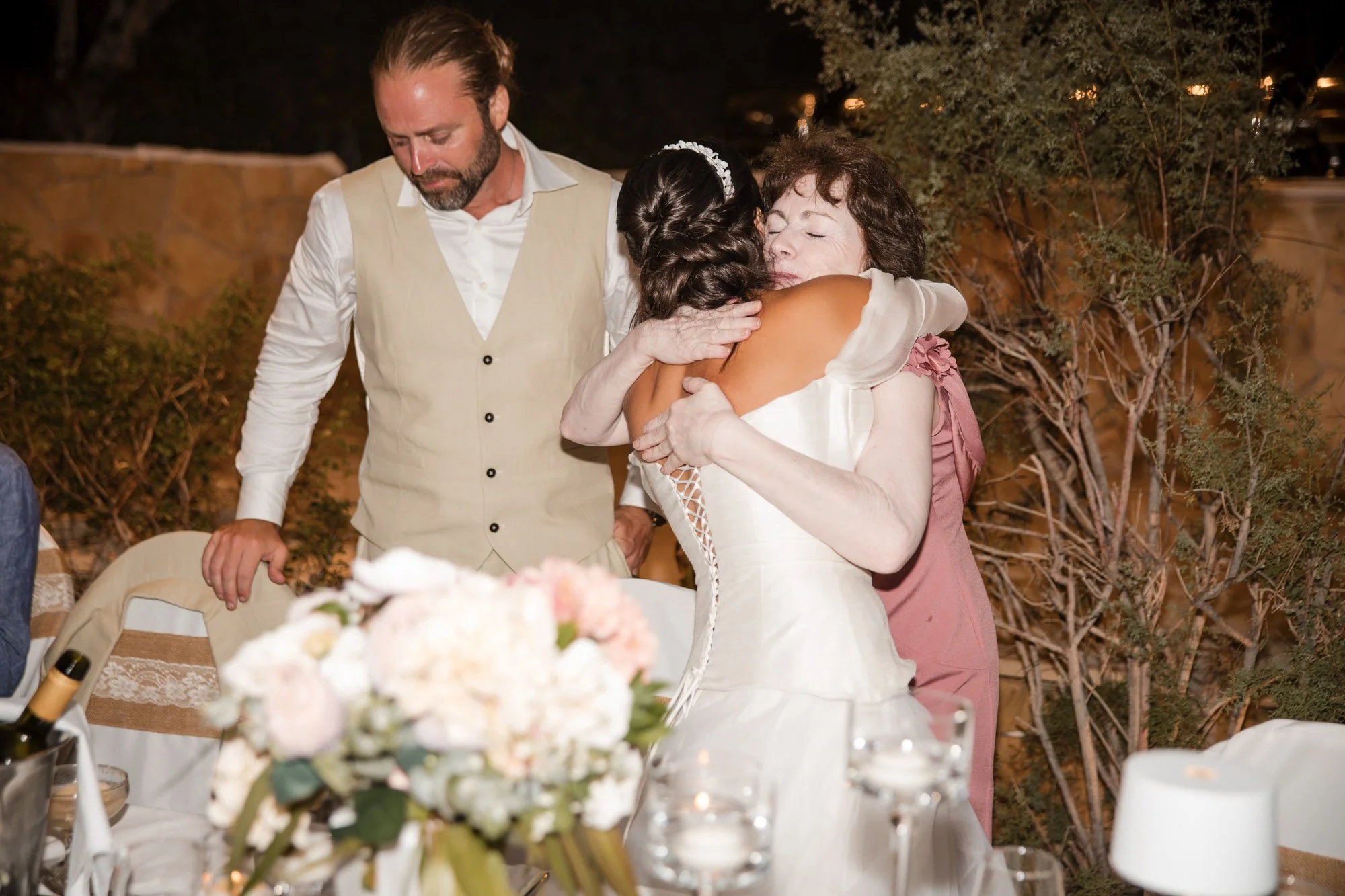 Two women hugging each other with emotional expressions, one with dark hair in a white dress and another with curly brown hair in a mauve dress, while a man in a beige vest and white shirt stands nearby at a restaurant table decorated with flowers an