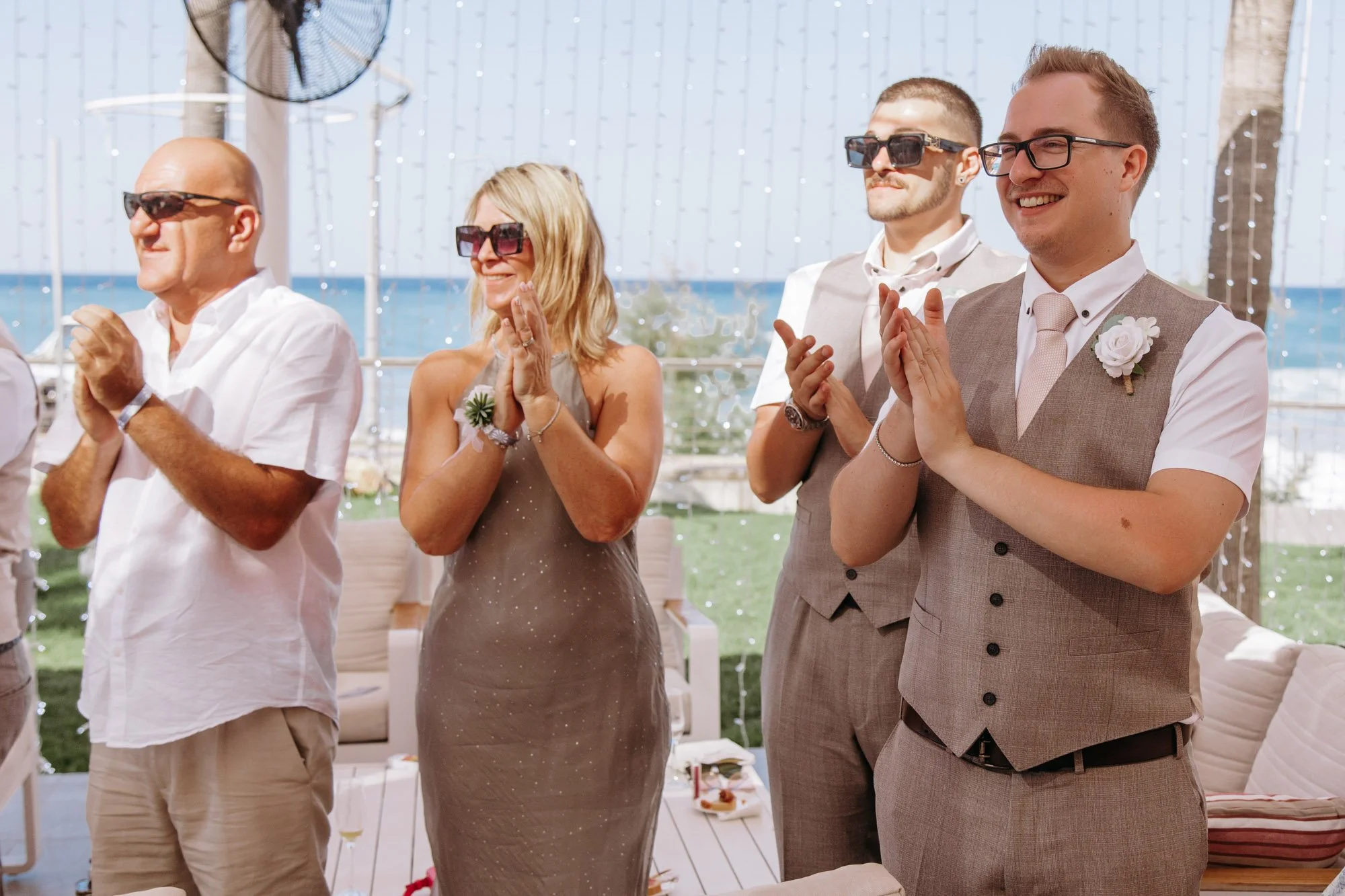 Four people standing outdoors, clapping and smiling, at a wedding celebration near the beach with string lights overhead.