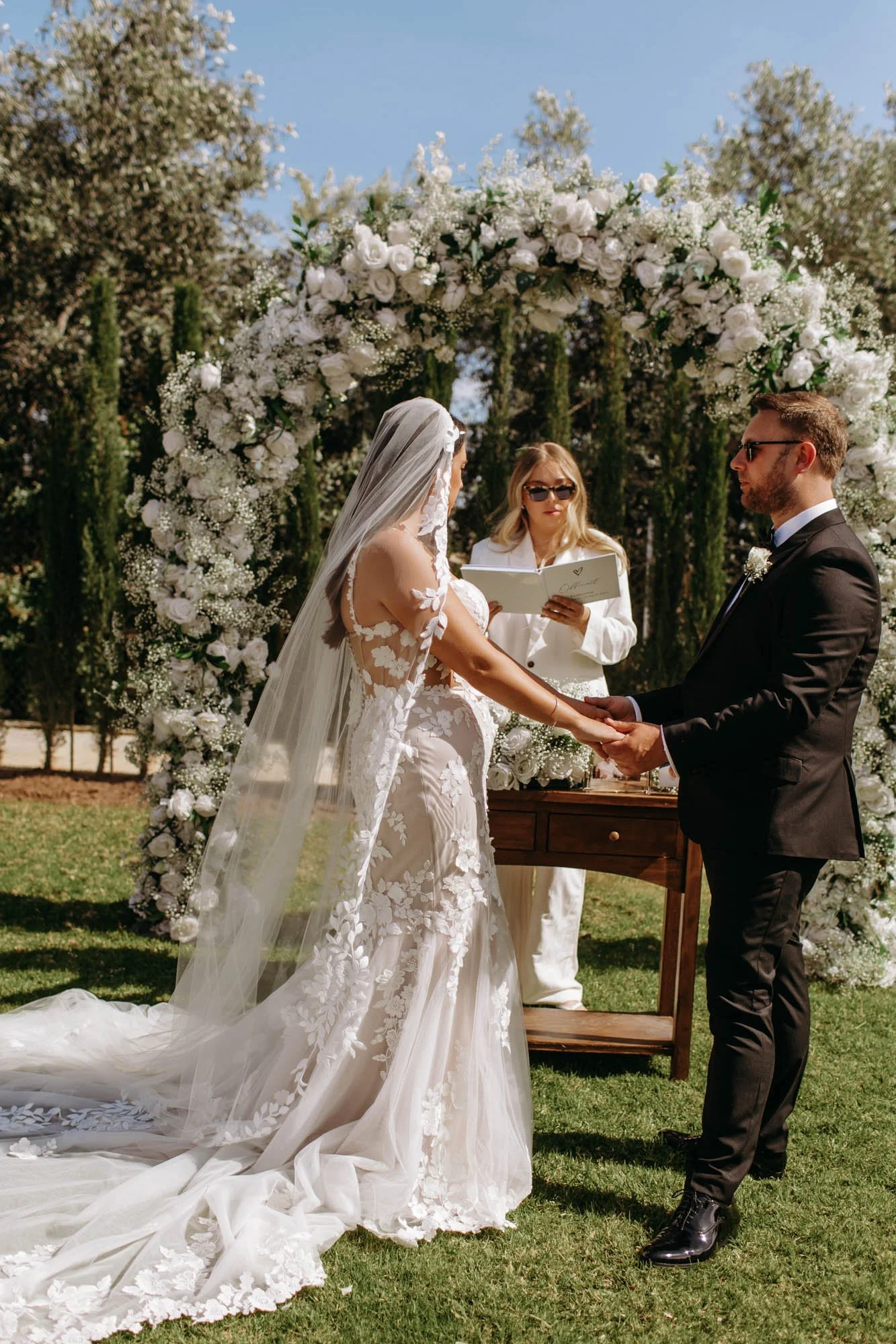 A couple gets married outdoors under a floral arch with an officiant standing behind them. The bride is in a white, floral lace wedding gown with a veil, and the groom is in a black suit with sunglasses, holding hands during the ceremony.