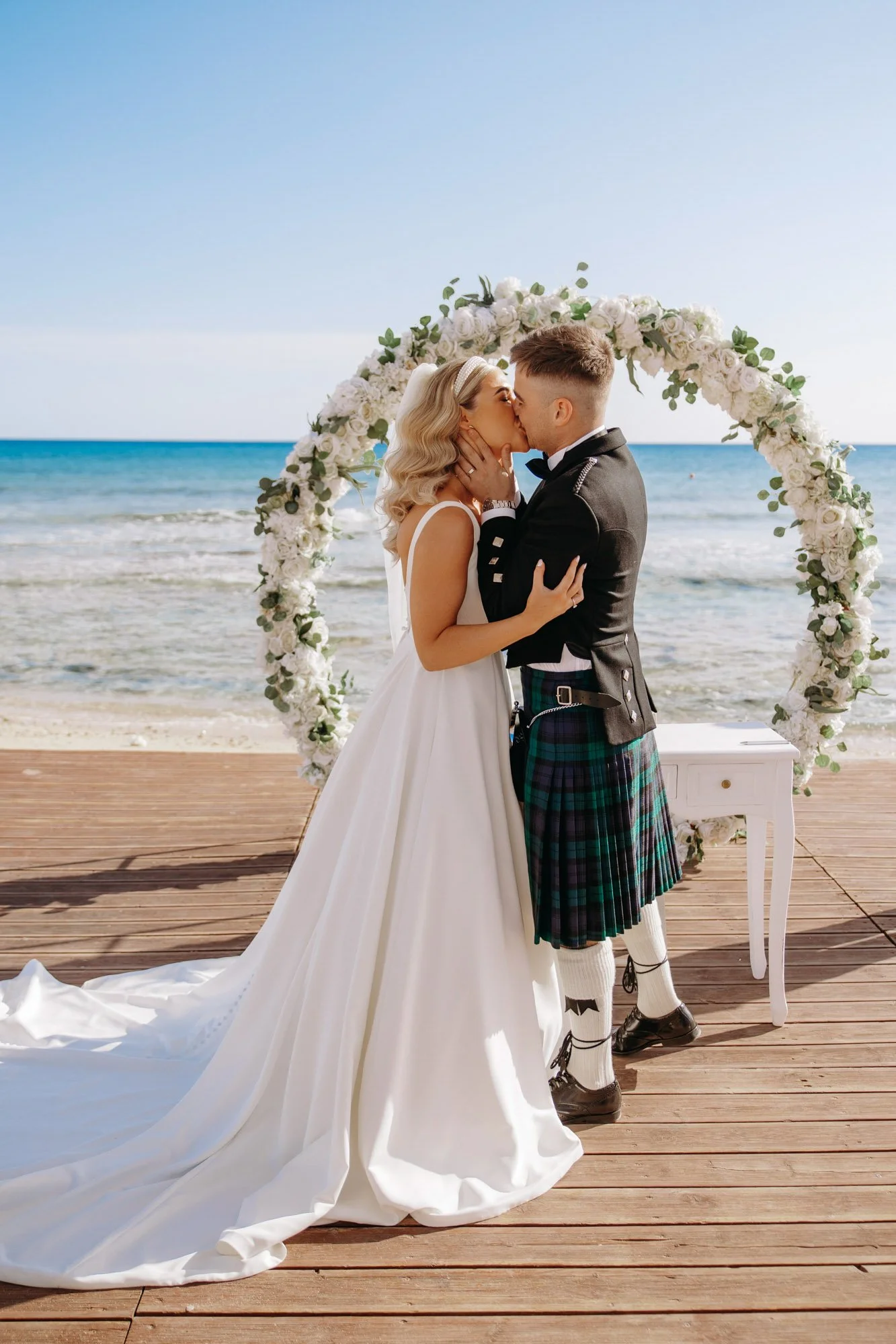 A couple in wedding attire sharing a kiss on a beach during their wedding ceremony, with a floral arch and ocean in the background.