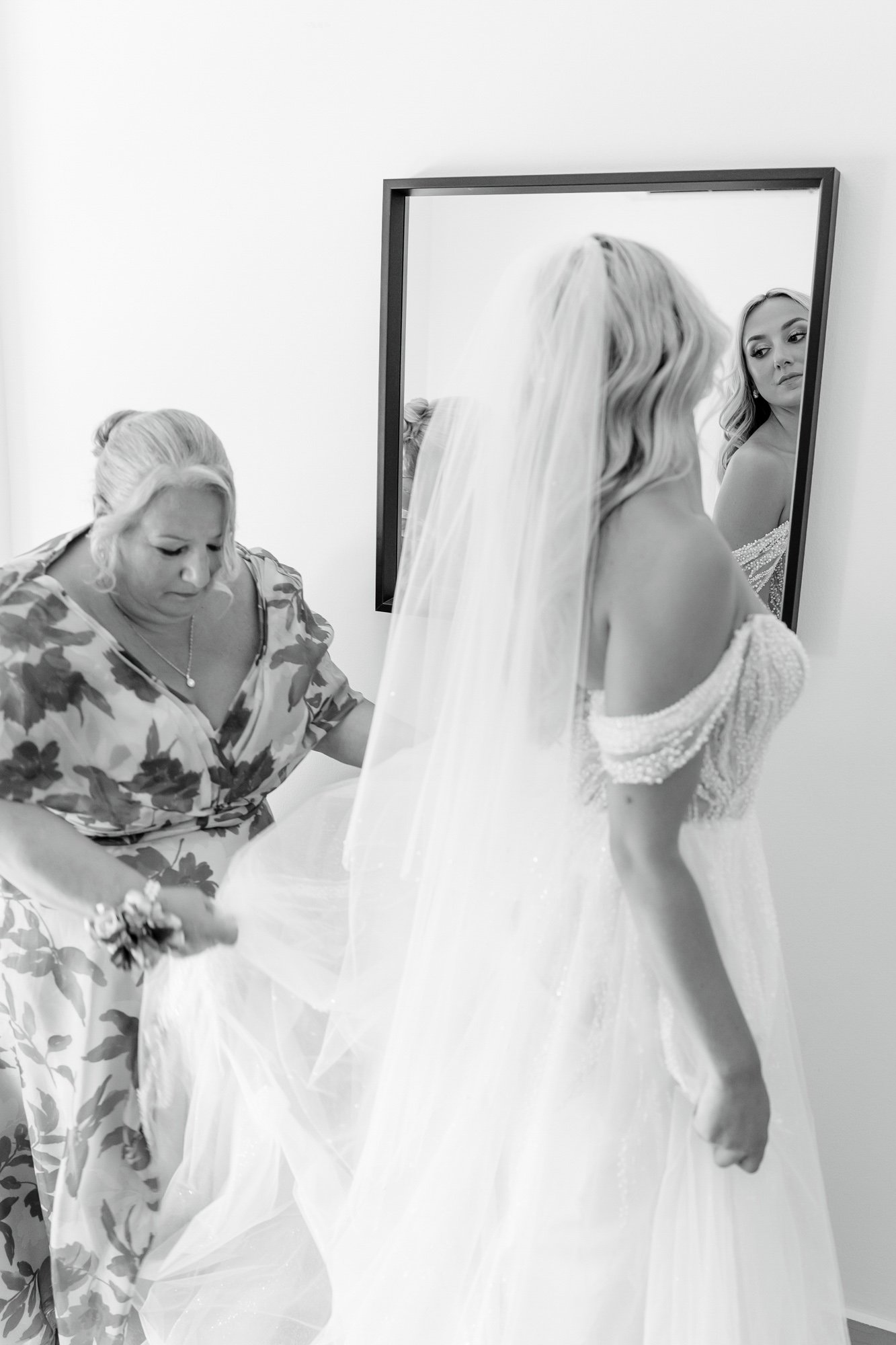 A bride in a wedding dress and veil standing in front of a mirror, with a woman helping her adjust the dress or veil, in a room with plain white walls.