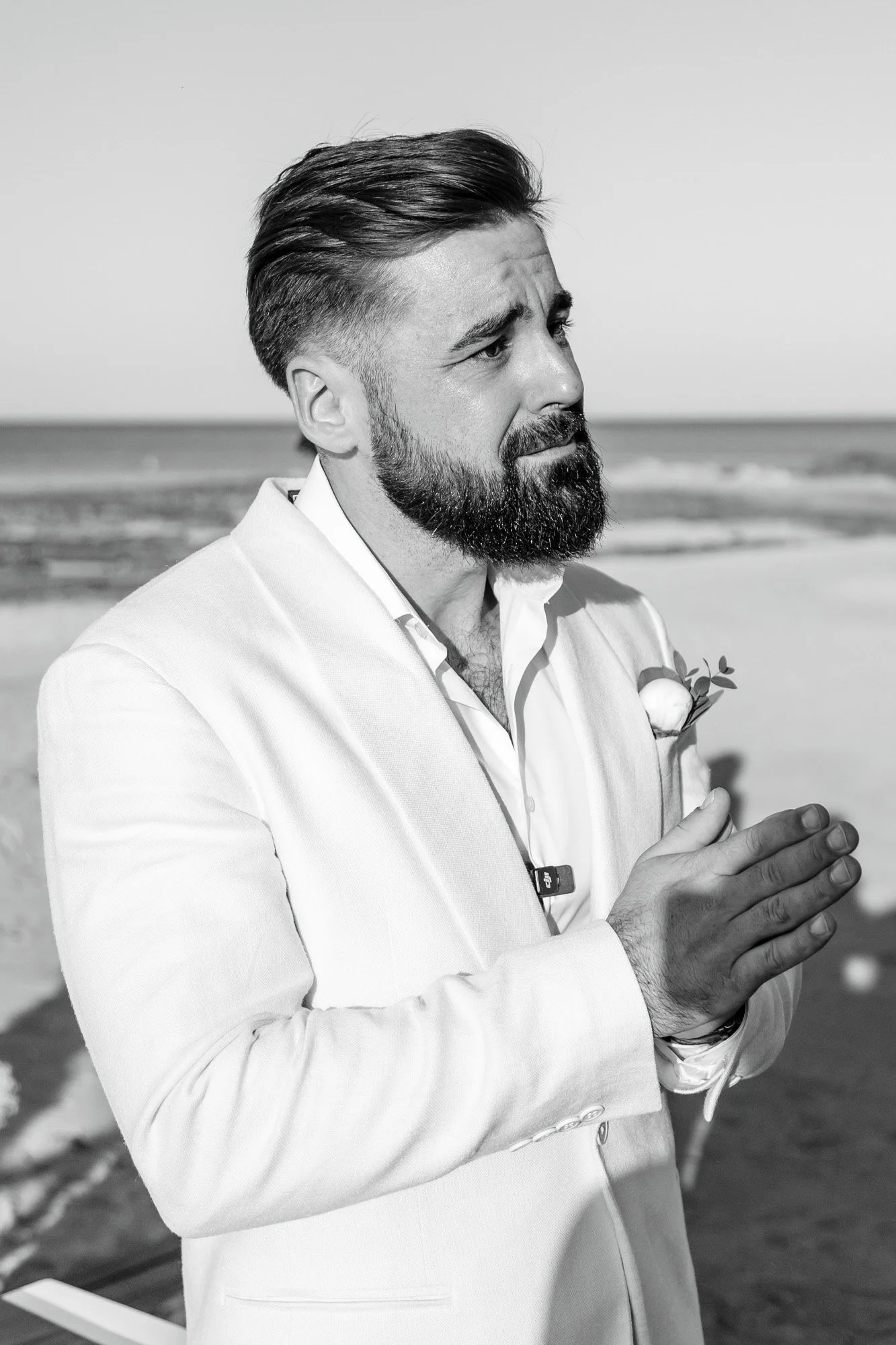 A bearded man in light suit with hands together on a beach, looking contemplative, black and white photograph.