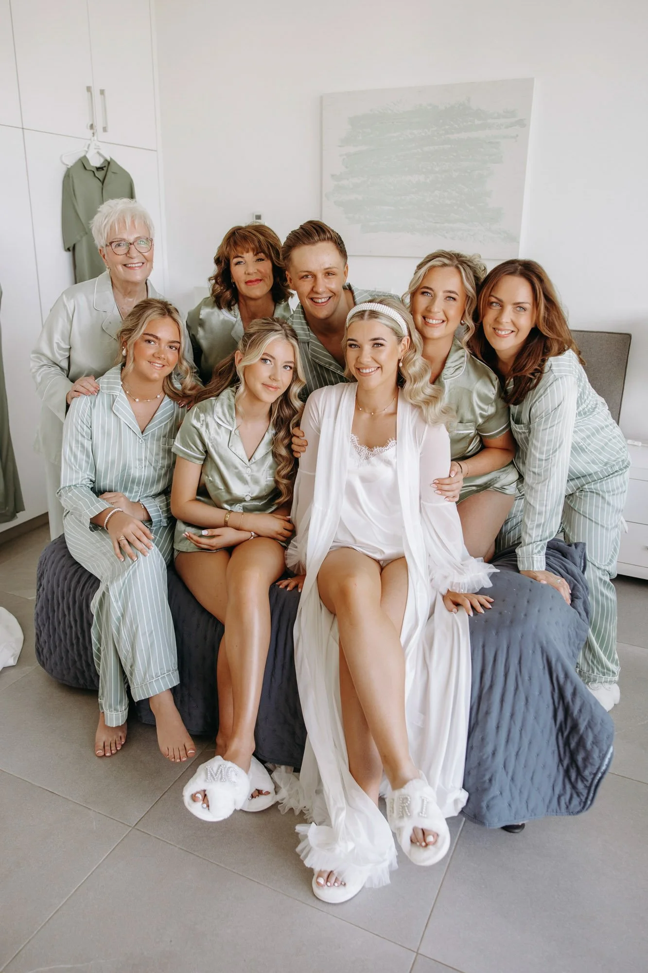 A bride and her bridesmaids sitting and standing around her on a bed, all dressed in pajamas, smiling in a bright, modern bedroom.