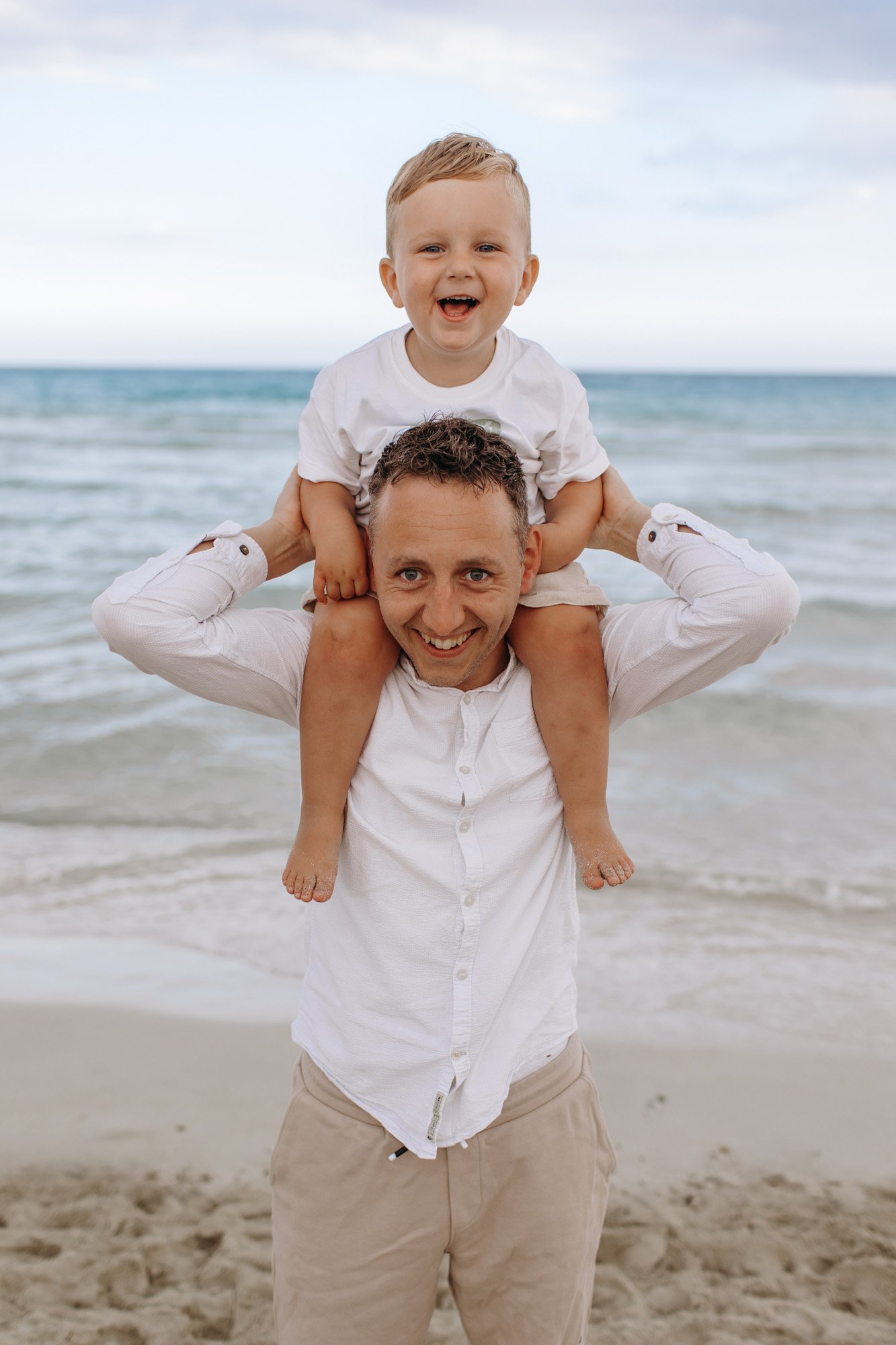 A man holding a young boy on his shoulders at the beach, both smiling, with the ocean and cloudy sky in the background.