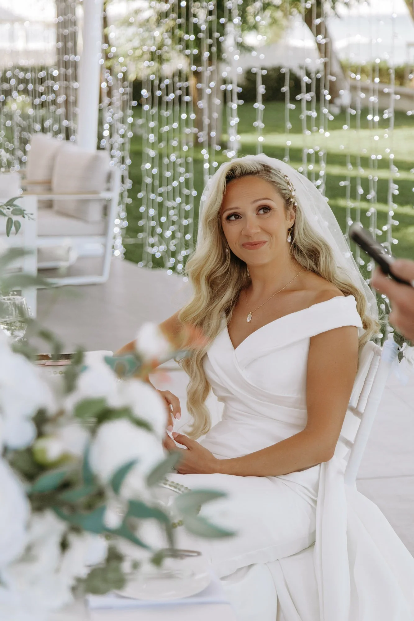 A bride with long blonde hair in a white wedding gown, sitting outdoors with fairy lights in the background, smiling softly.