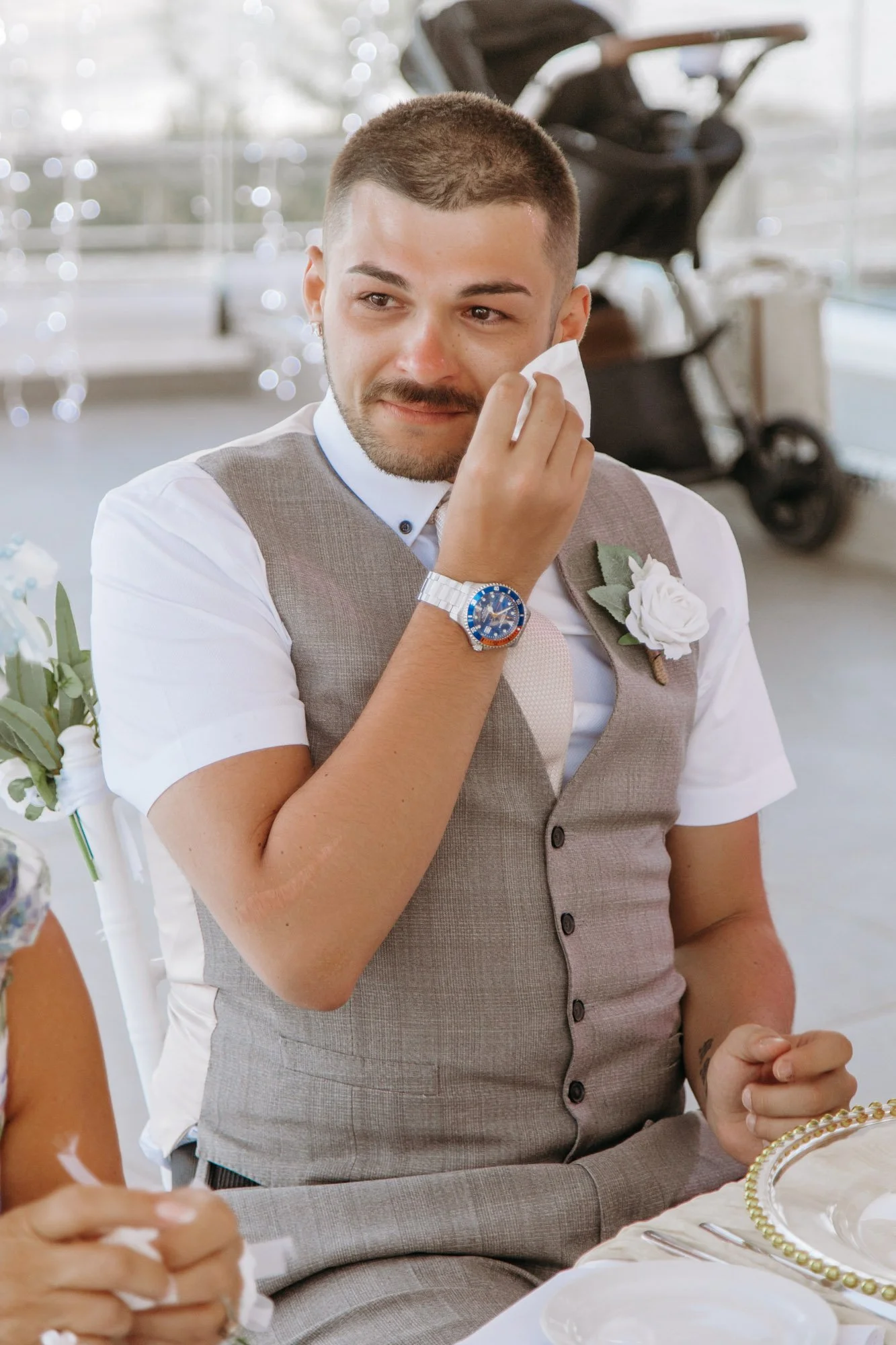 A young man with short hair, a beard, and a mustache, wiping his eye with a tissue at a formal event. He is wearing a gray vest, white shirt, and a watch, with a white flower pinned to his vest.