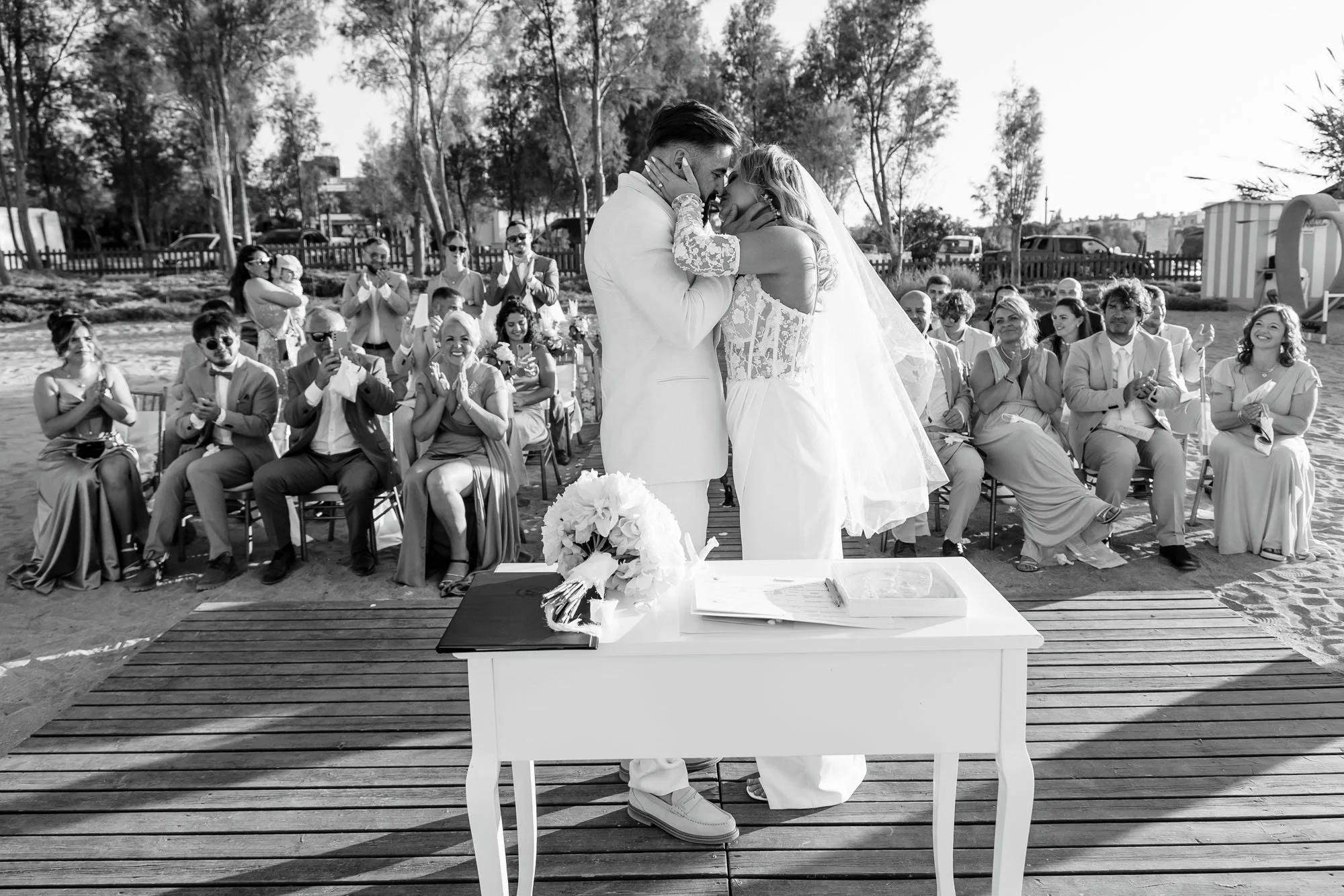 A black and white photo of a wedding ceremony on a beach, with a couple kissing at the altar while friends and family clap and smile in the background.