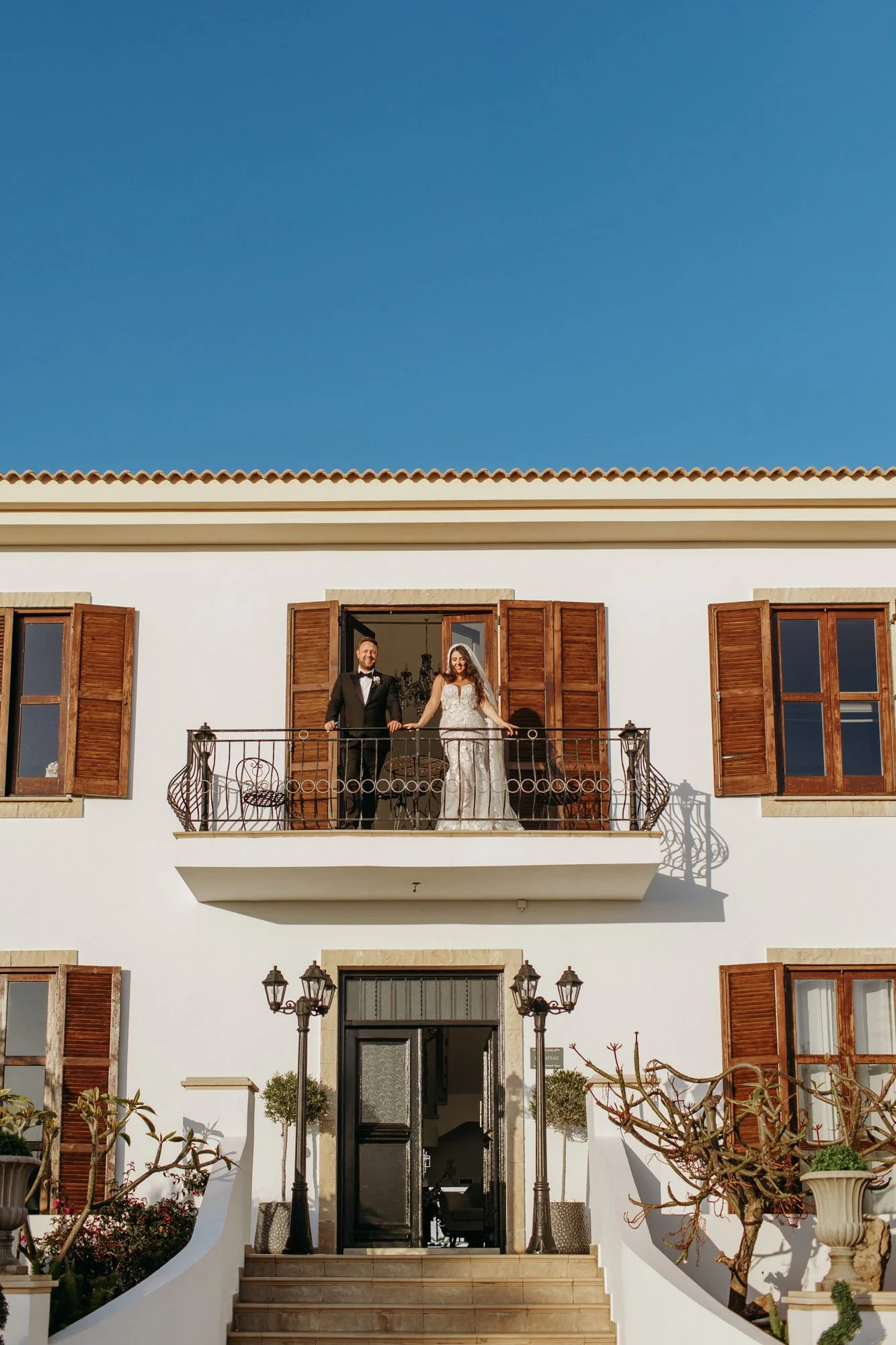 Couple in formal attire standing on a balcony of a white building with wooden shutters and outdoor lanterns, under a clear blue sky.