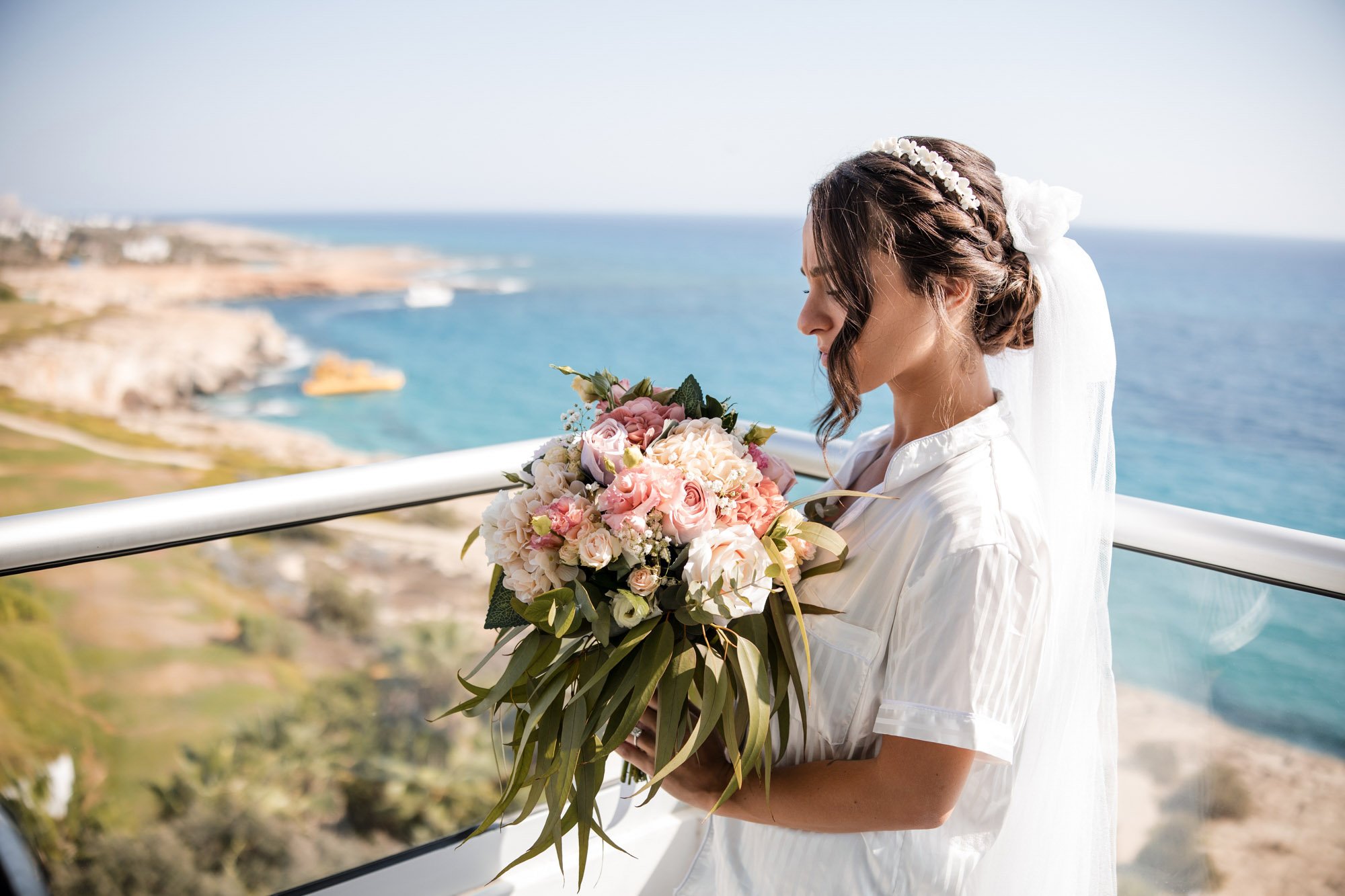 A bride in a white dress holding a bouquet of pink and white flowers, standing on a balcony with a coastal view of the ocean and cliffs in the background.