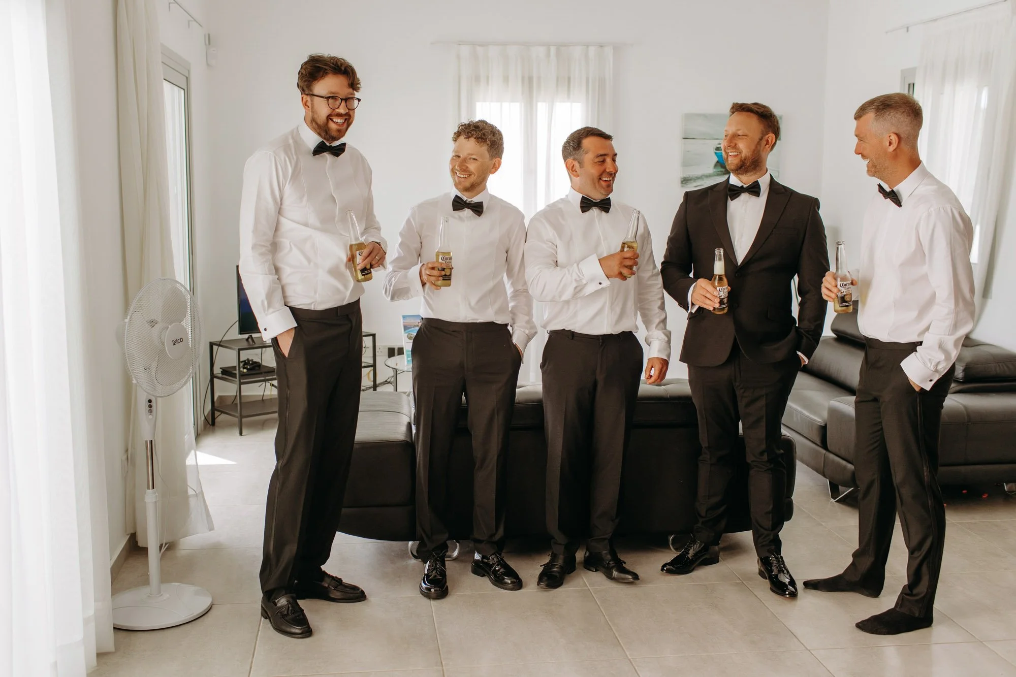 Six men dressed in formal attire, five in white shirts with black bow ties and one in a black suit with a black bow tie, standing and holding bottles of beer in a bright, modern living room, engaging in conversation and smiling.