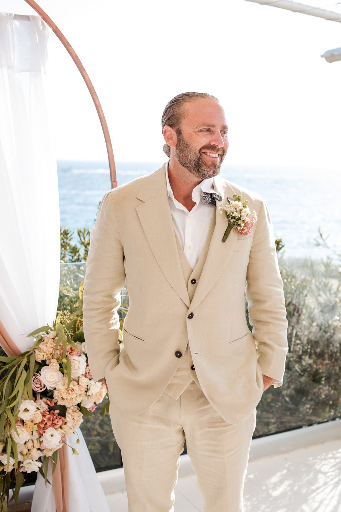 A man in a cream-colored suit with a white shirt and boutonniere, standing outdoors by the ocean with a floral arrangement nearby, smiling.