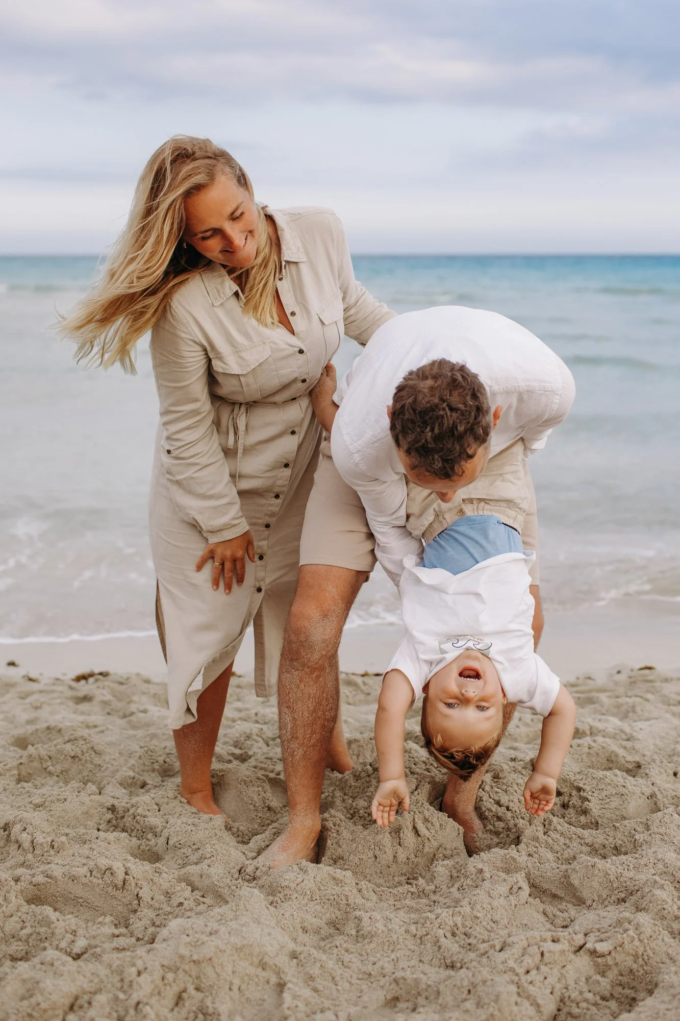 A woman and two children playing on the beach. The woman is smiling and looking down at the children, one of whom is upside down with a big smile, while the other is on her hands and knees in the sand.