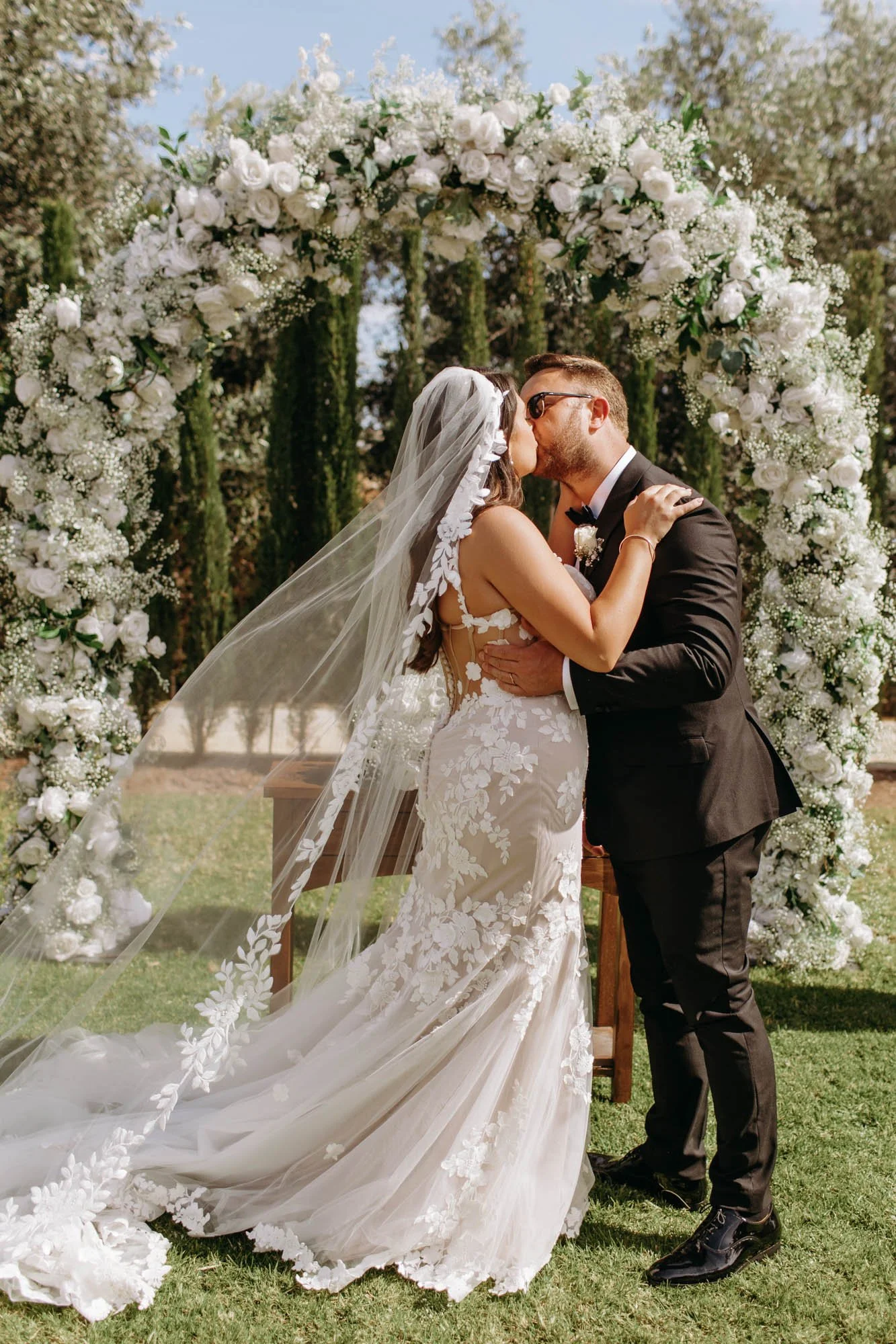 A bride and groom share a kiss in front of a large floral arch during their outdoor wedding ceremony.
