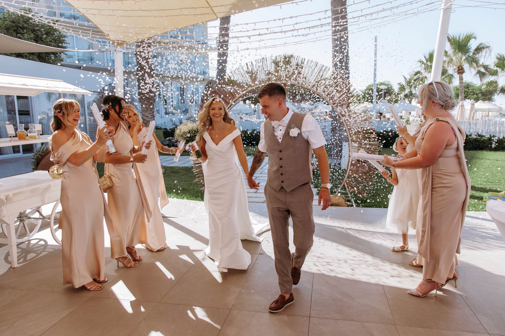 A bride and groom holding hands walking through a celebration circle with confetti falling, surrounded by smiling bridesmaids and guests at an outdoor wedding reception with palm trees and white tents.