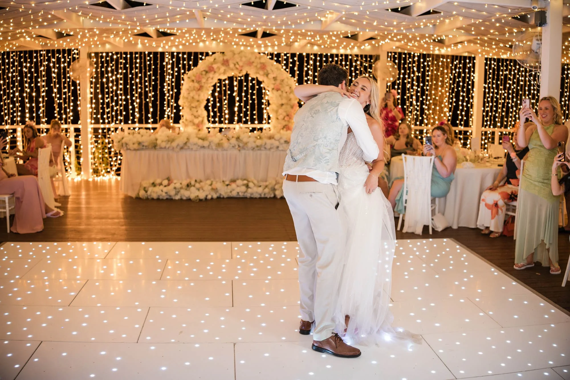 A bride and groom dancing at their wedding reception with guests watching and taking photos, decorated with fairy lights and a floral arch in the background.