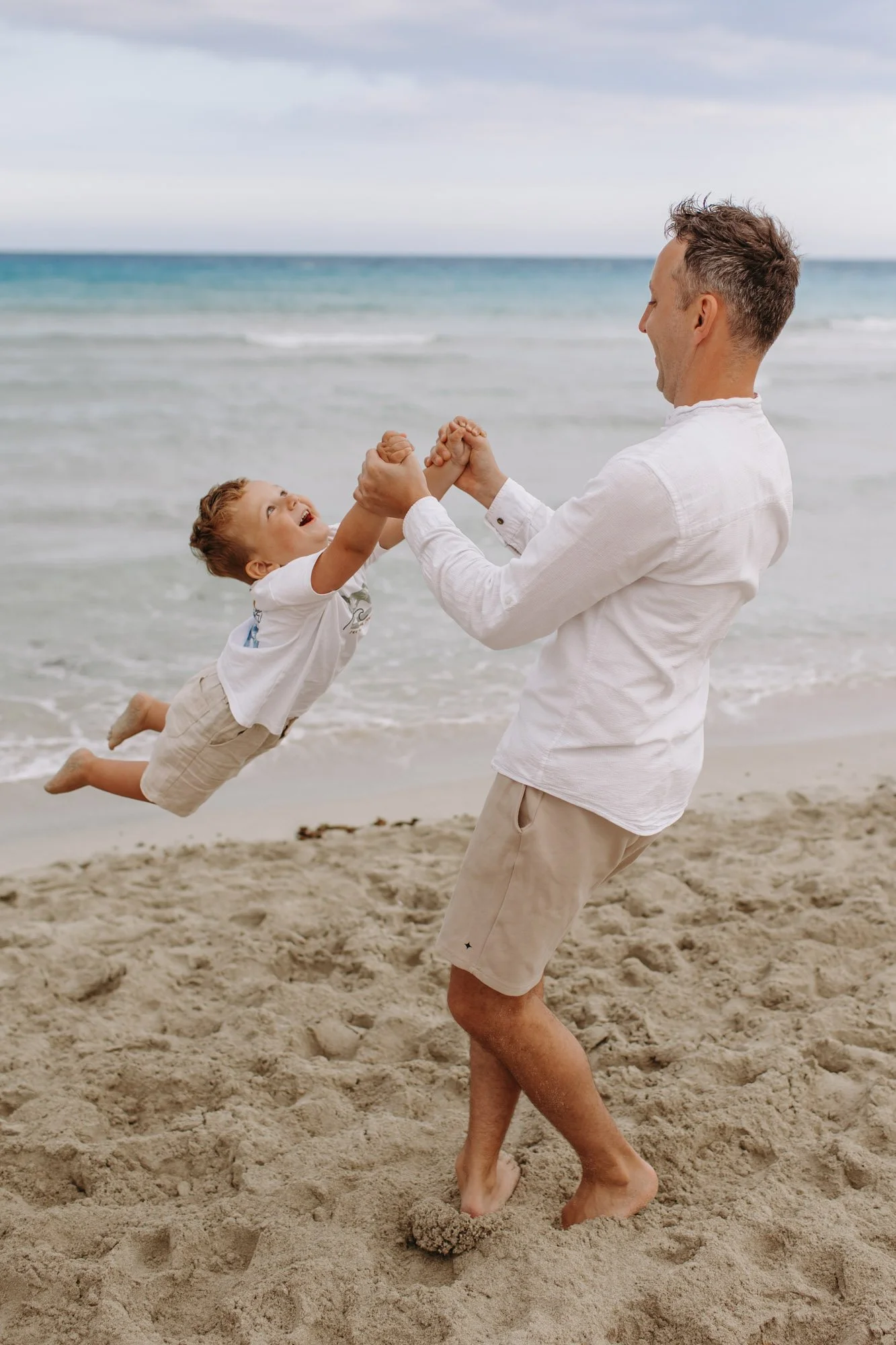 A man and a young boy playing on the beach, the boy is jumping and holding the man's hands, sand and ocean in the background.