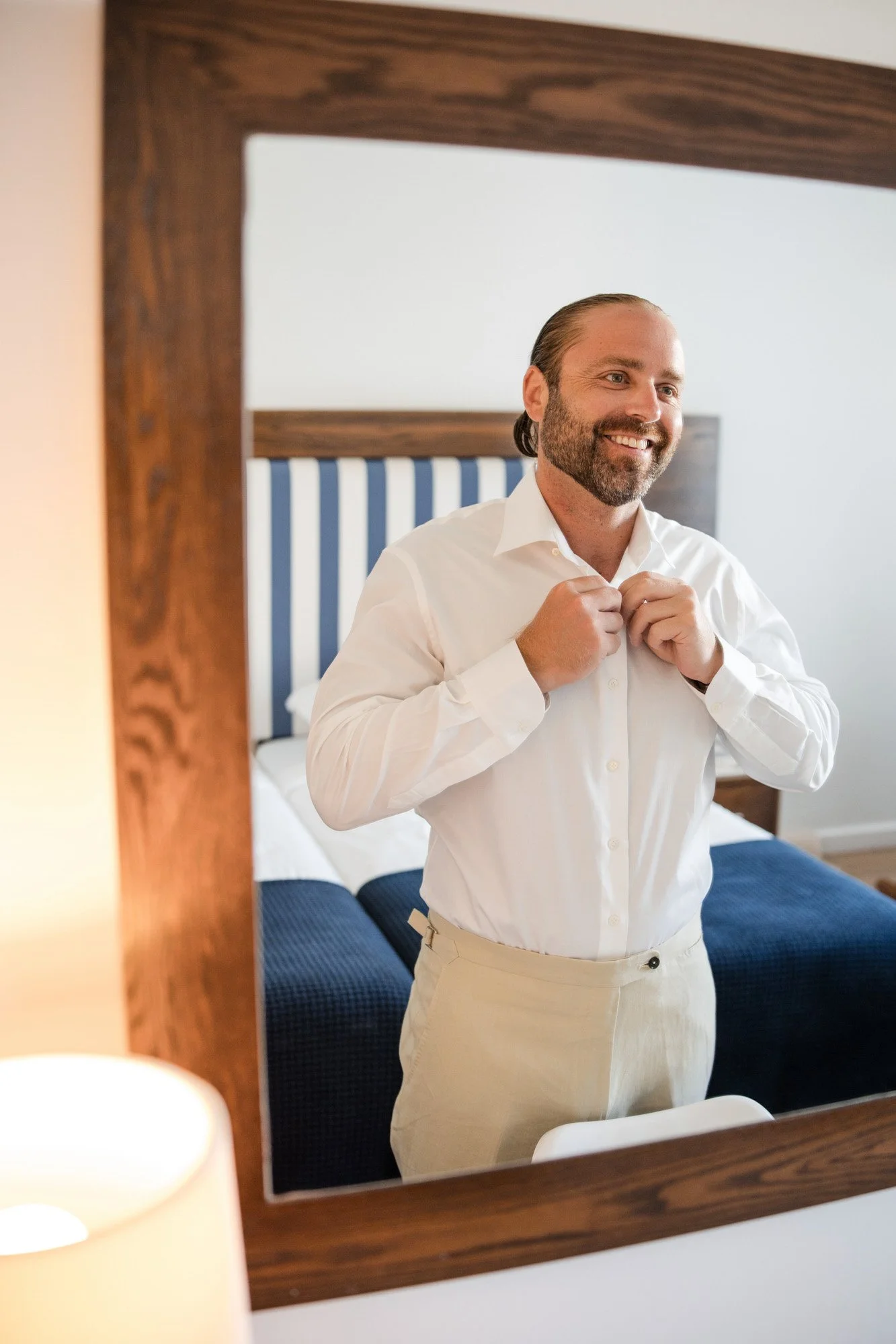 A man with a beard and slicked-back hair standing in front of a mirror, buttoning his white shirt, smiling, in a bedroom with a bed that has a blue and white striped headboard.