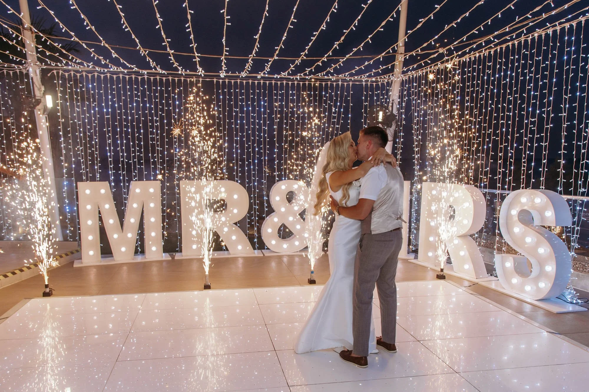 A newlywed couple sharing a kiss on a dance floor at night, with large illuminated letters spelling 'MR & R'S' behind them. The backdrop features hanging string lights and sparkling fireworks.