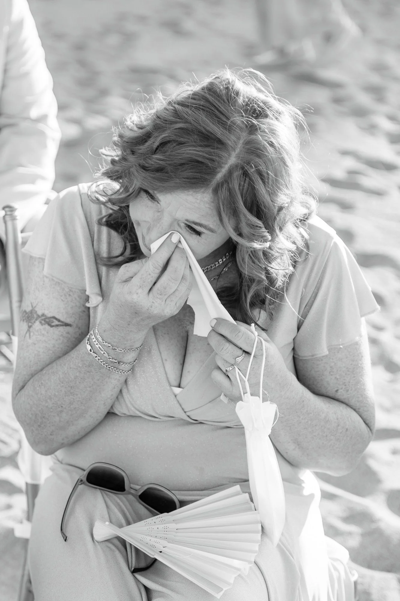A woman sitting outdoors on a beach, wiping away tears with a tissue, holding a face mask, with sunglasses and a fan on her lap, appearing emotional.
